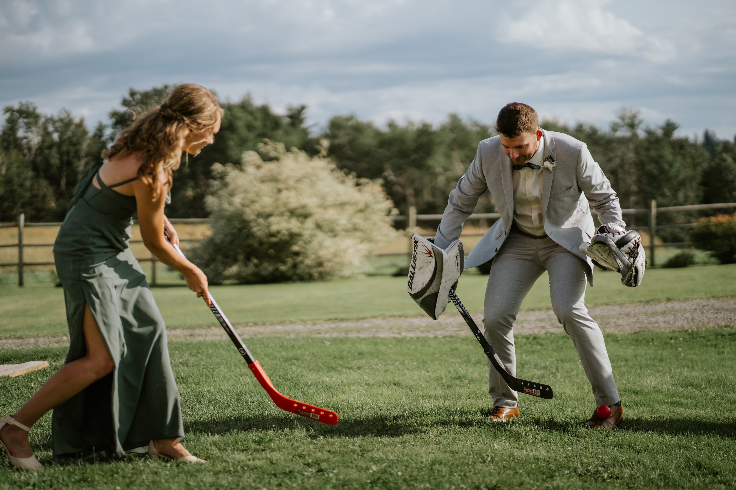 A woman and a man playing hockey on a grassy field outdoors, with trees and a fence in the background.