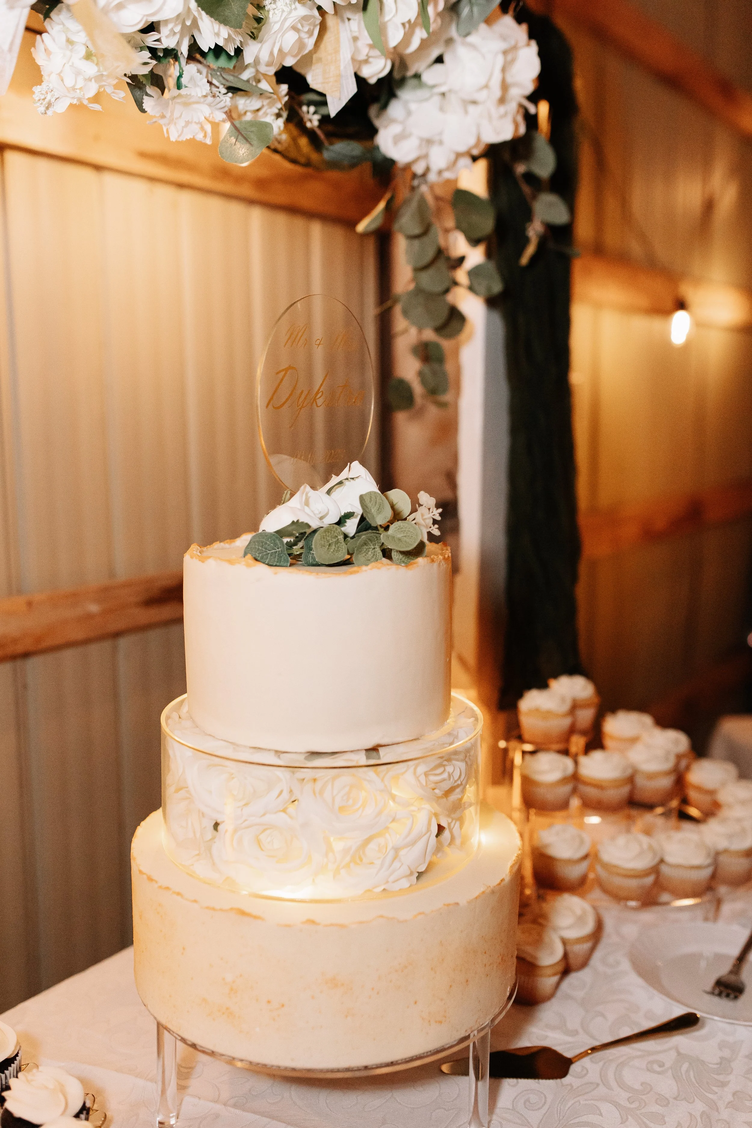A three-tiered wedding cake decorated with white frosting, rose-shaped decorations, and greenery, with a clear topper featuring the words 'Mr & Mrs Dykstra.' The cake is displayed on a stand, with cupcakes in the background and floral arrangements ab