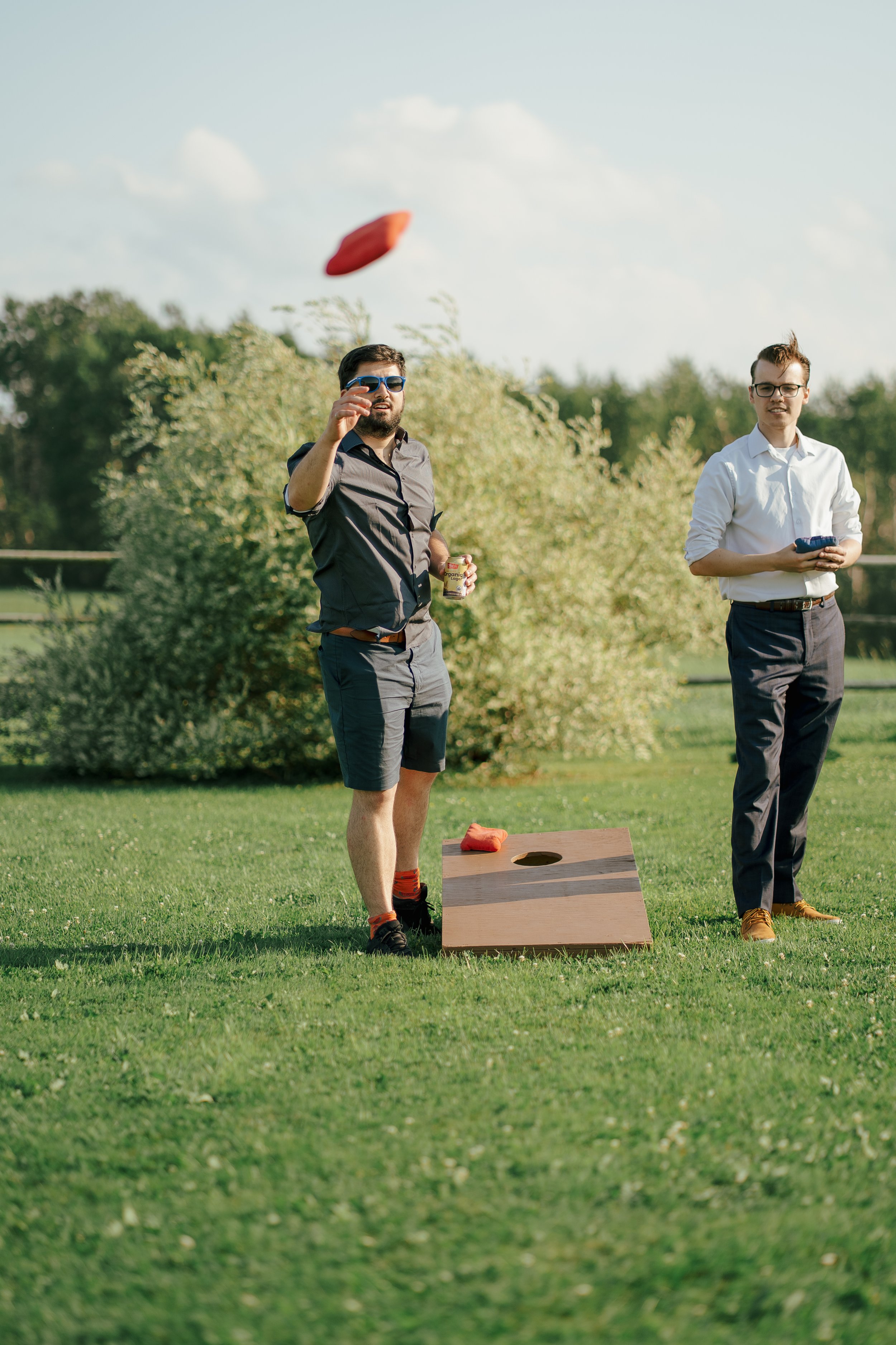 Two men playing cornhole outdoors on a grassy field, one tossing a bean bag while the other holds a device.