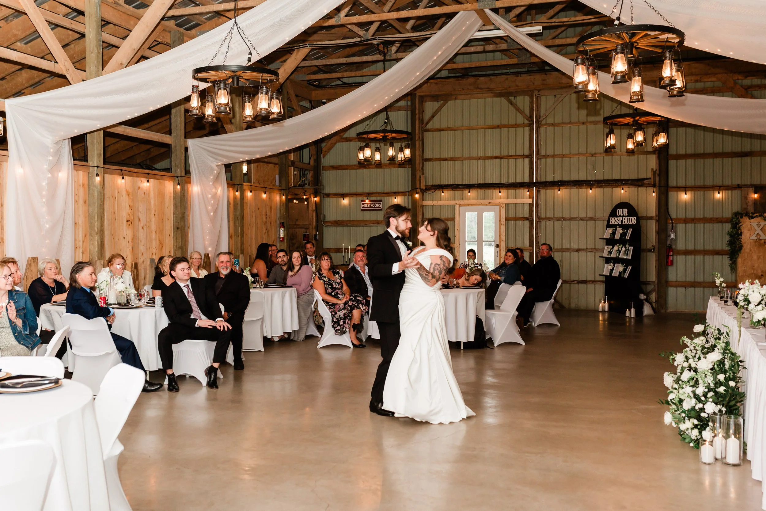 A bride and groom dancing at their wedding reception in a barn with wooden walls and ceiling, decorated with string lights and draped fabric. Guests seated at tables watch the couple dance.