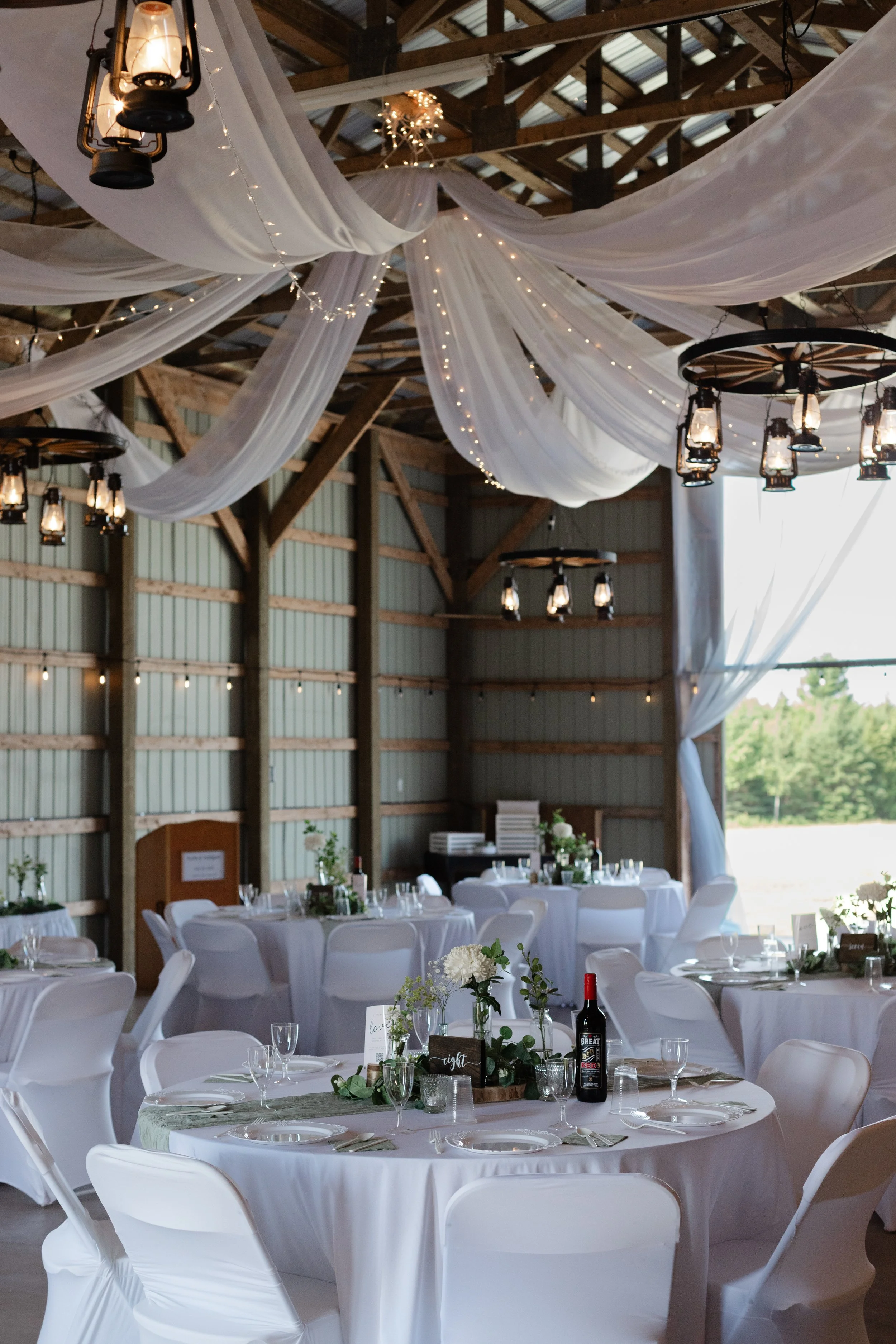 Indoor wedding reception decorated with white drapes, hanging lanterns, string lights, and round tables with white tablecloths and floral centerpieces.