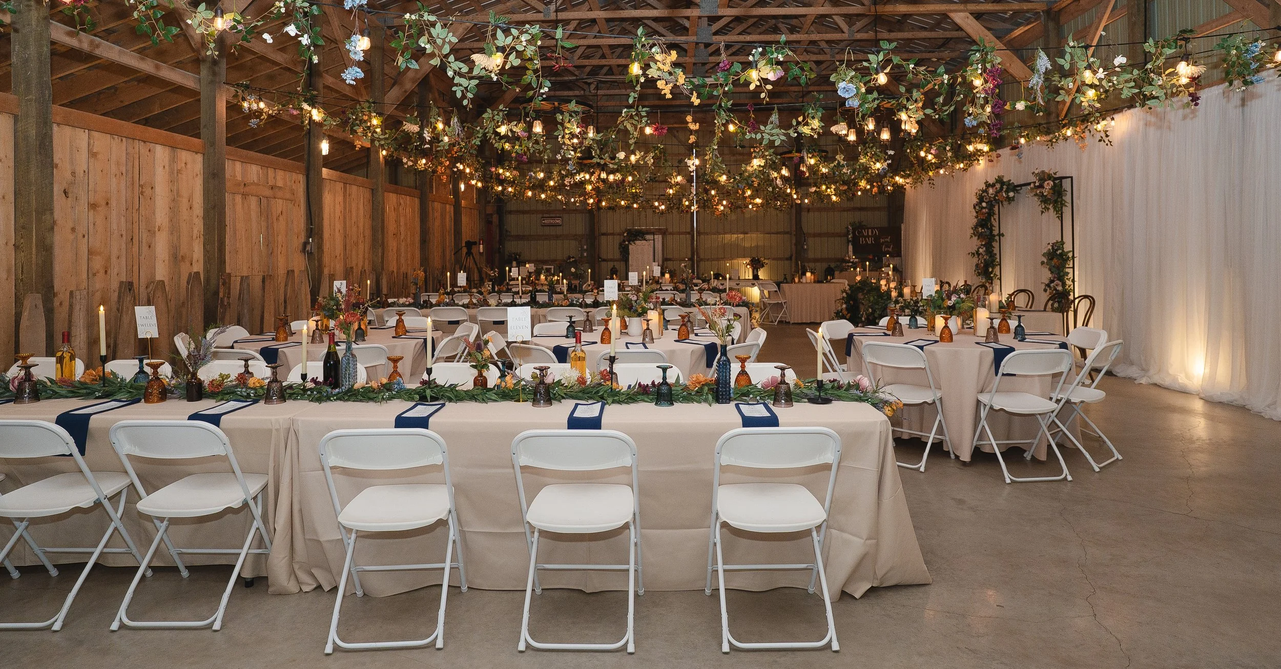 Wedding reception setup in a rustic barn with long tables decorated with greenery, candles, and flowers. Ceiling is adorned with hanging lights and floral arrangements. White curtains and wooden wall panels in the background.