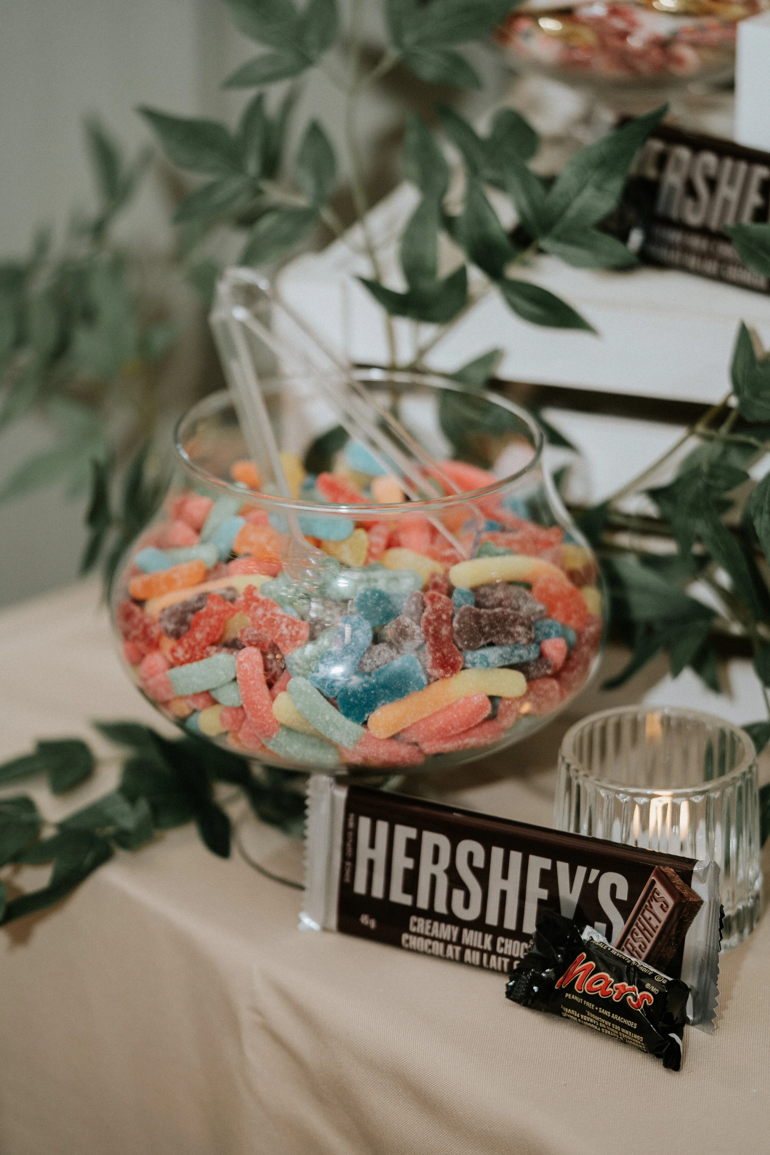 A glass bowl filled with colorful gummy candies, surrounded by a Hershey's chocolate bar and a Mars chocolate bar on a table decorated with green leaves.
