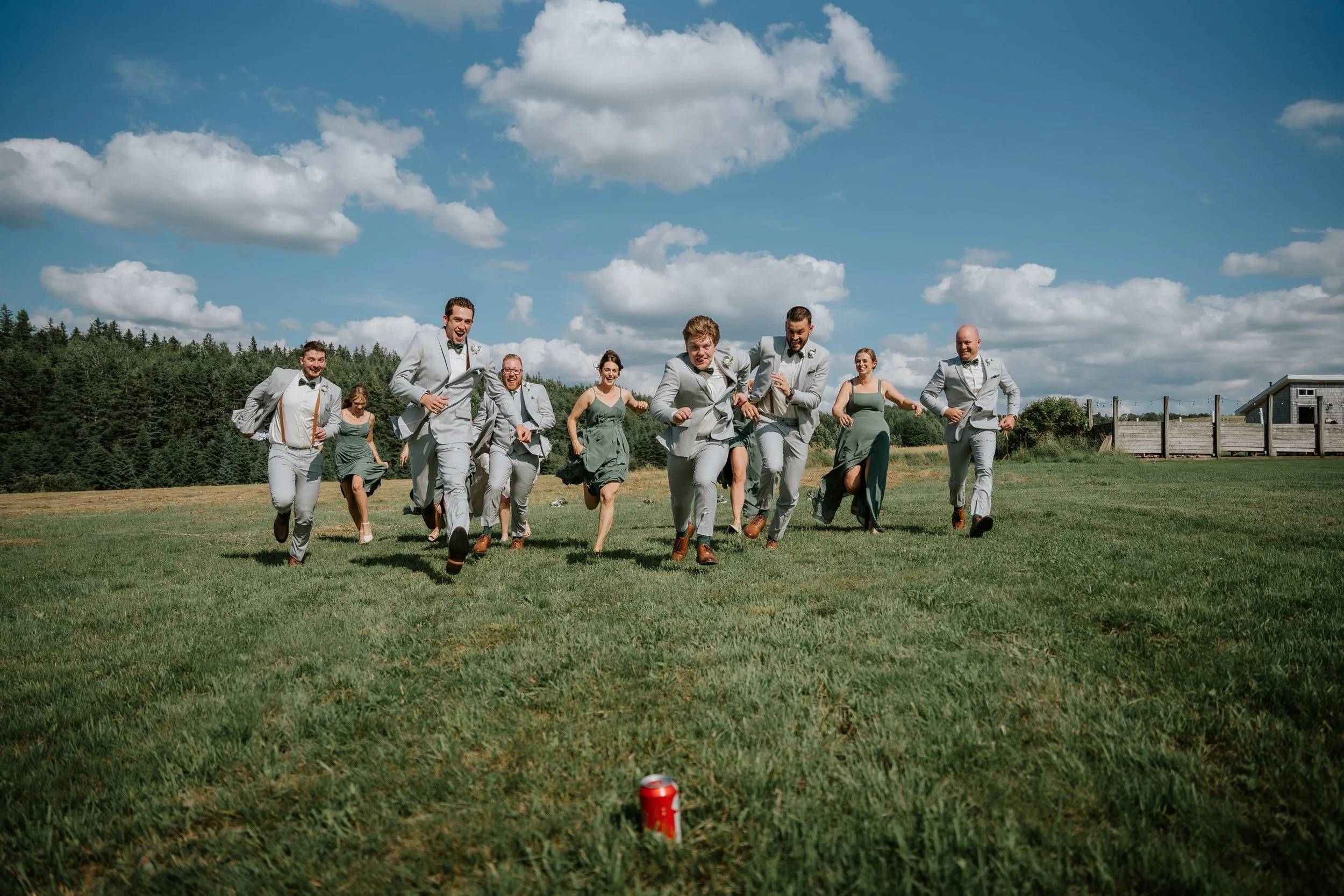 A group of people in formal attire running towards a red soda can on grass outdoors on a sunny day.