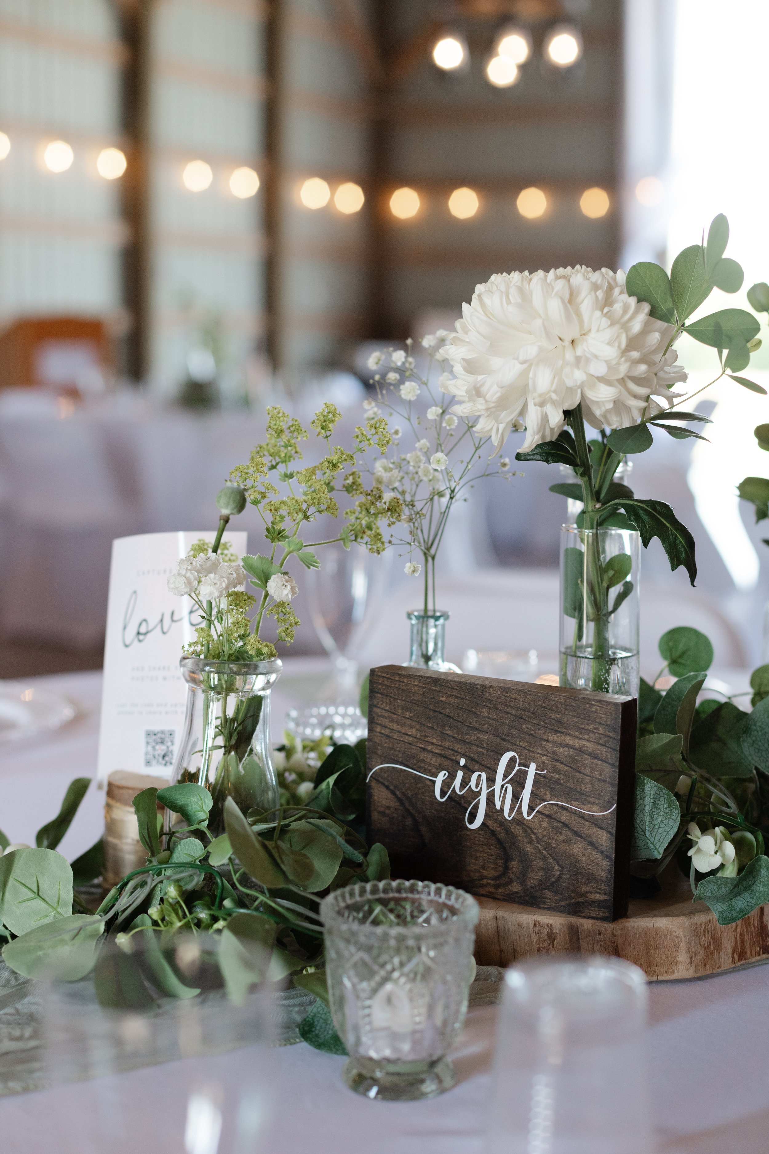 Decorative table centerpiece with white flowers, glass vases, greenery, a wooden 'eight' table number sign, and blurred background of a rustic event space with string lights.