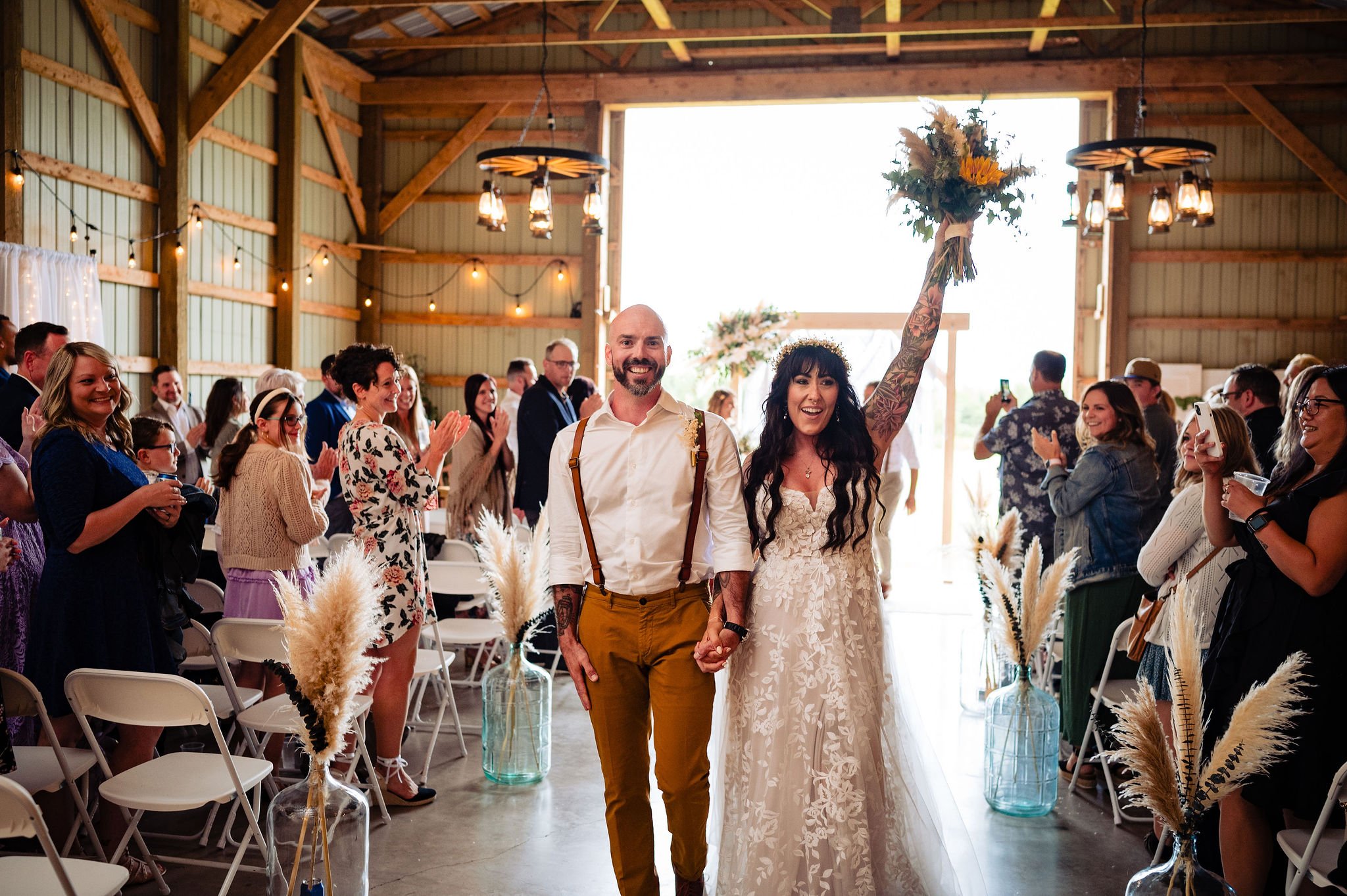 A bride and groom walking hand in hand down the aisle after their wedding ceremony, smiling and holding a bouquet, as guests applaud in a rustic barn setting.