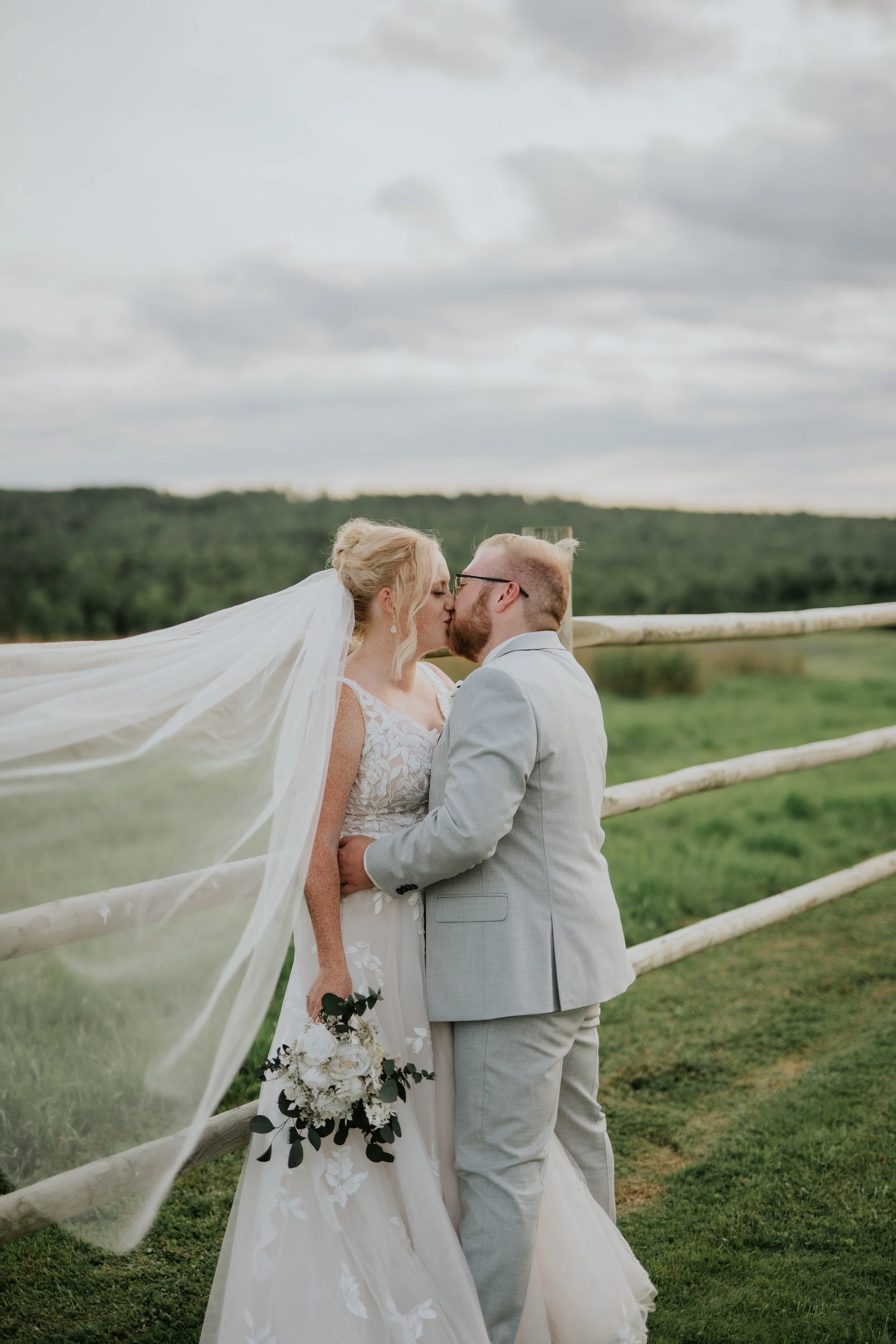 A bride and groom share a kiss outdoors on their wedding day, leaning against a wooden fence with a scenic green landscape and cloudy sky in the background.