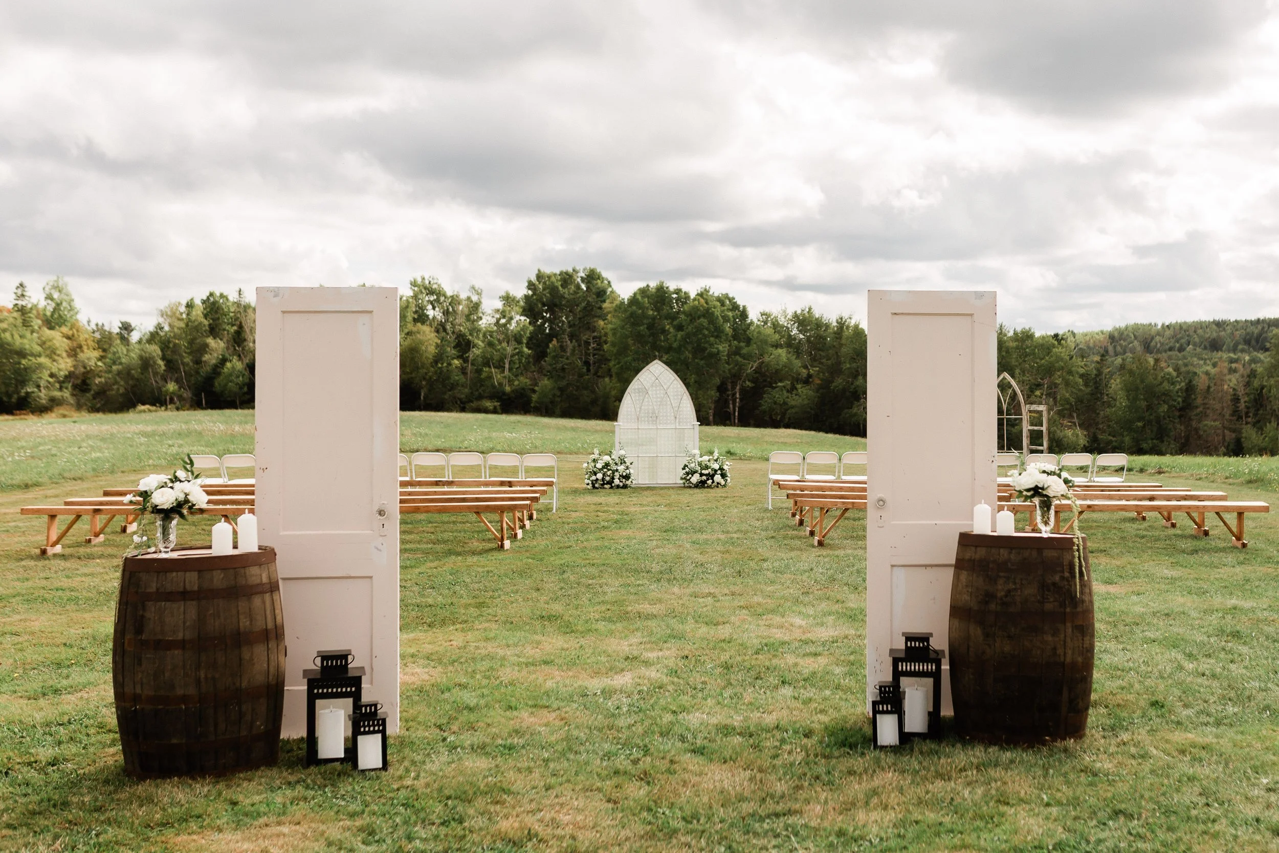 Outdoor wedding ceremony setup with wooden benches, white doors, floral arrangements, candles, and an arch in a green grassy field surrounded by trees.