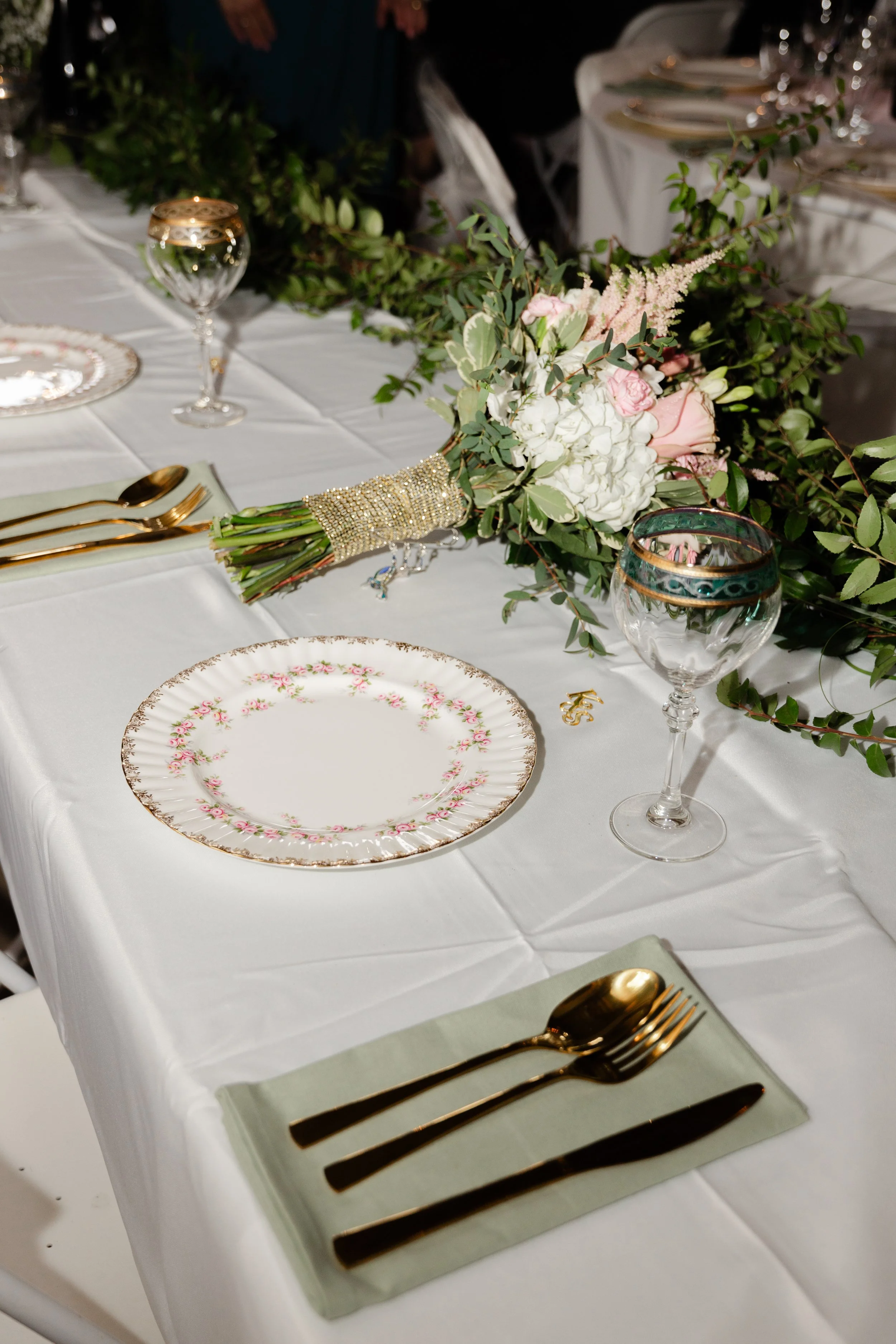 Formal dining table with a white tablecloth, floral centerpiece, gold cutlery, decorative plates, and wine glasses, set for a special occasion or wedding reception.