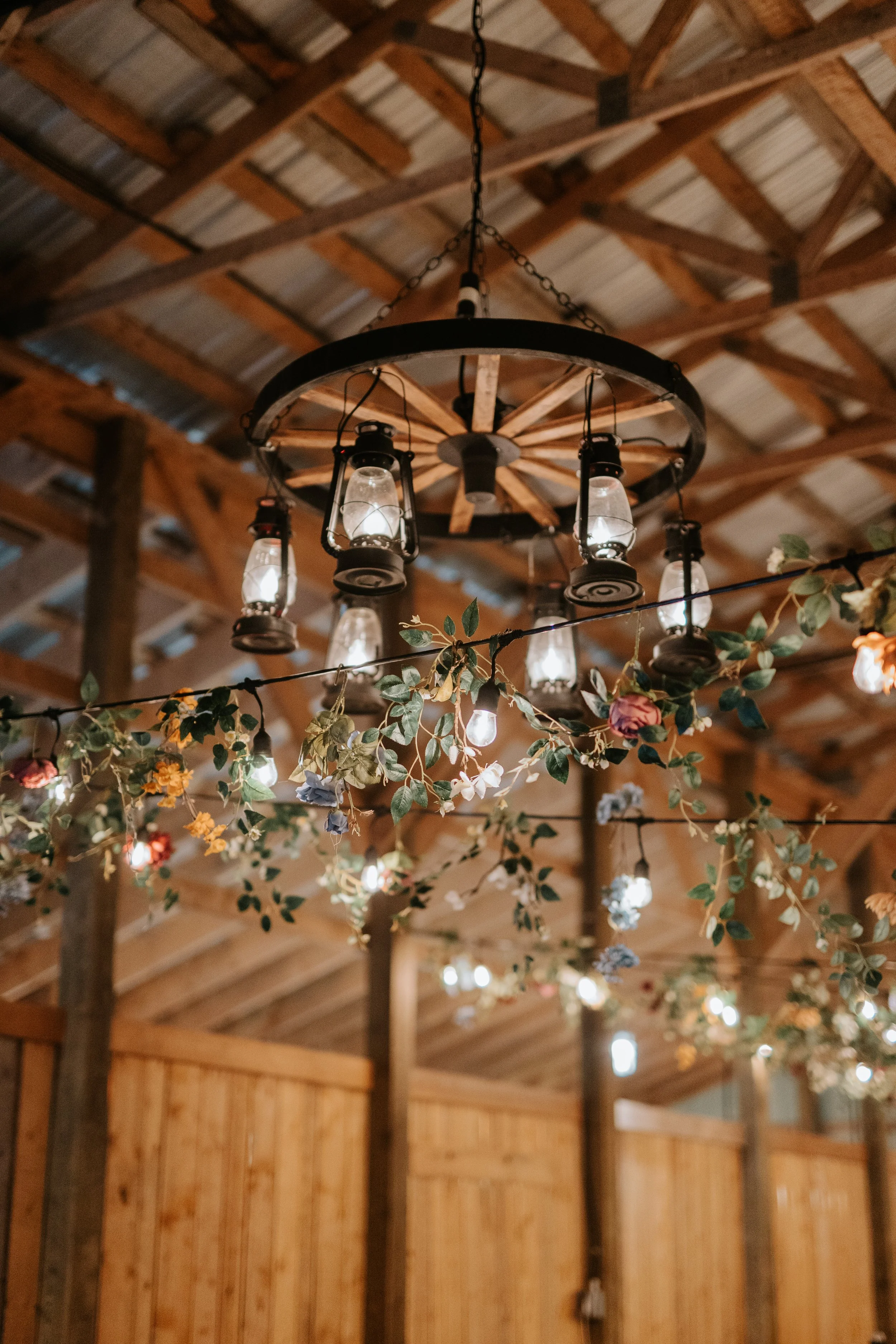 Chandelier with lantern-style lights hanging from a barn ceiling decorated with hanging floral garlands.