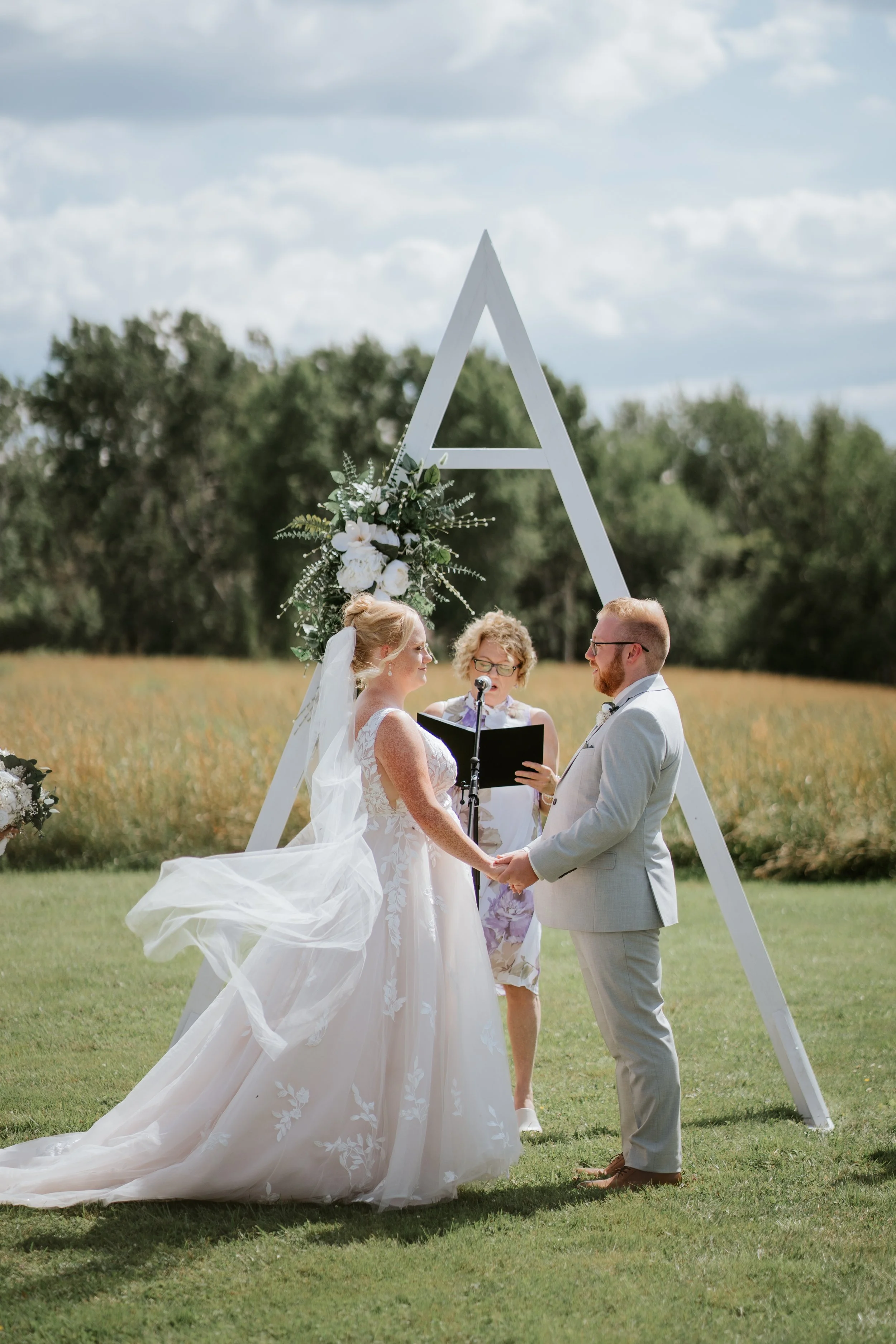 A couple gets married outdoors under a white triangular wedding arch decorated with white flowers and greenery, with a field and trees in the background.