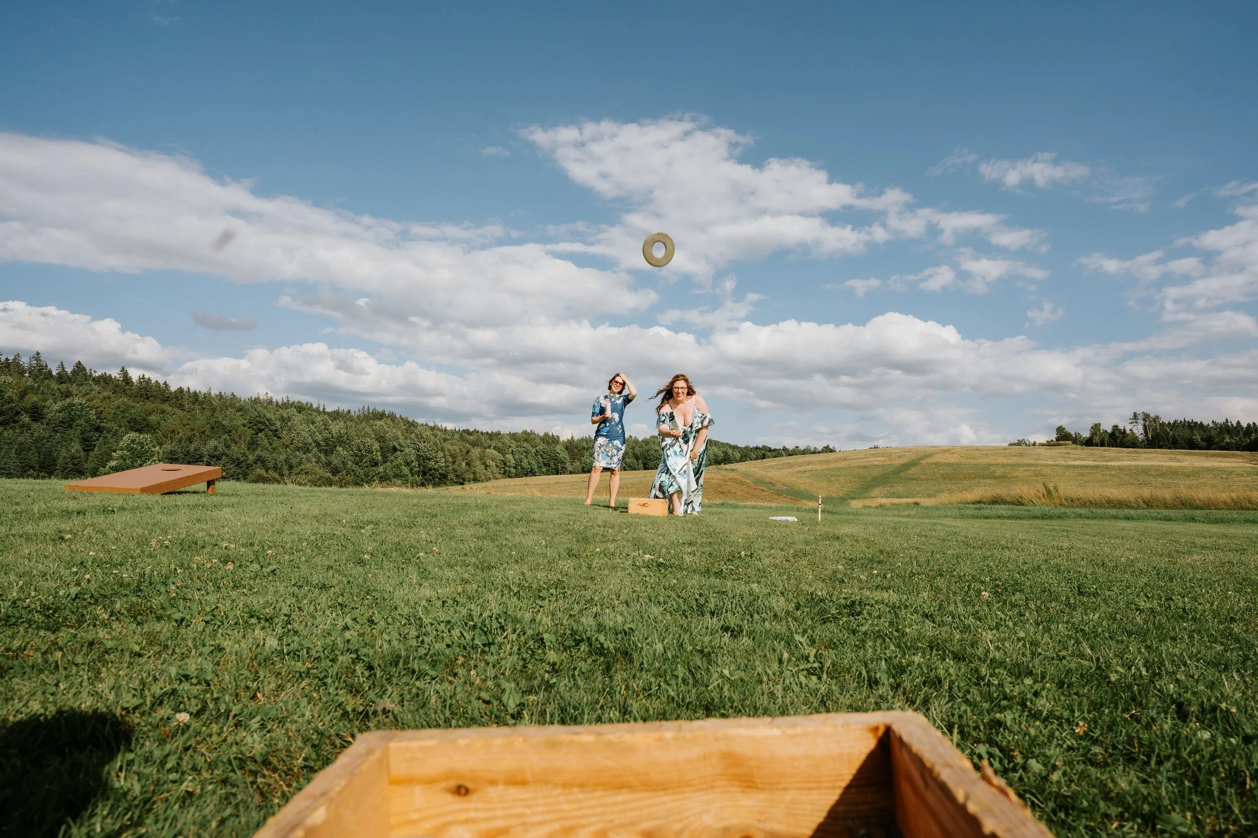 Two women in floral dresses are playing a game outdoors on a grassy field under a partly cloudy sky. One woman is about to throw a ring towards a target, while the other stands nearby. Wooden game pieces are visible in the foreground and background.