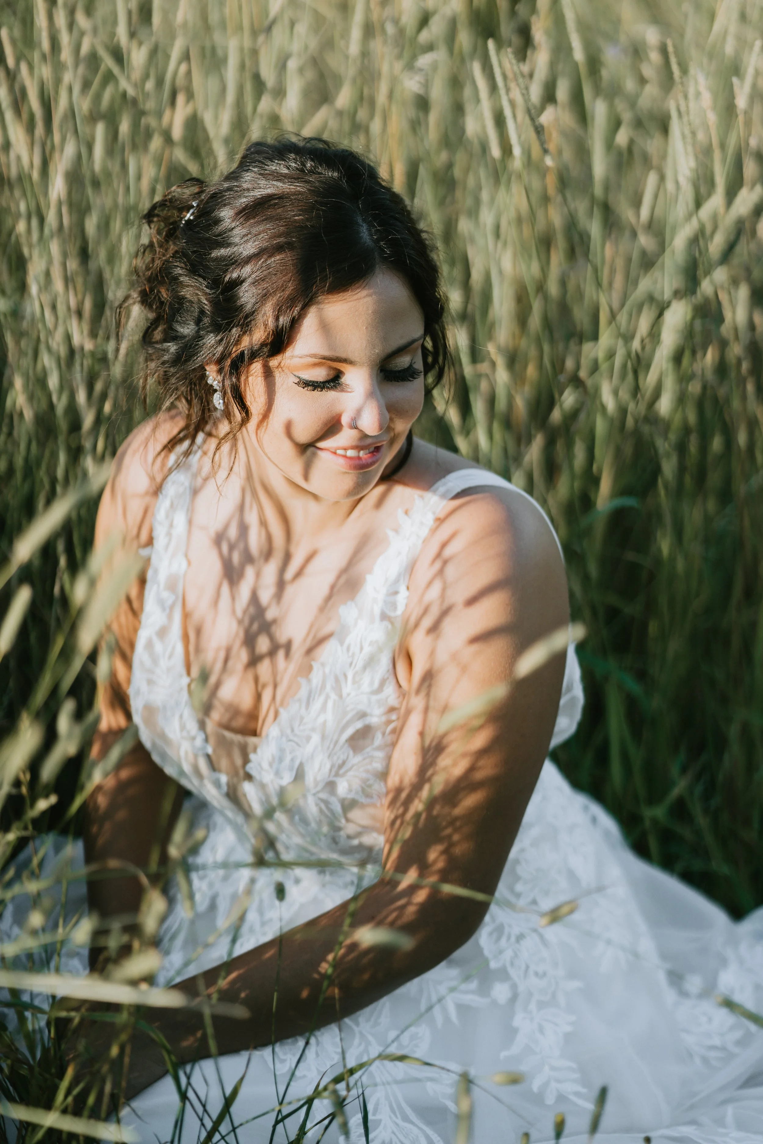 A woman in a white lace dress sitting in tall grass, smiling with eyes closed.