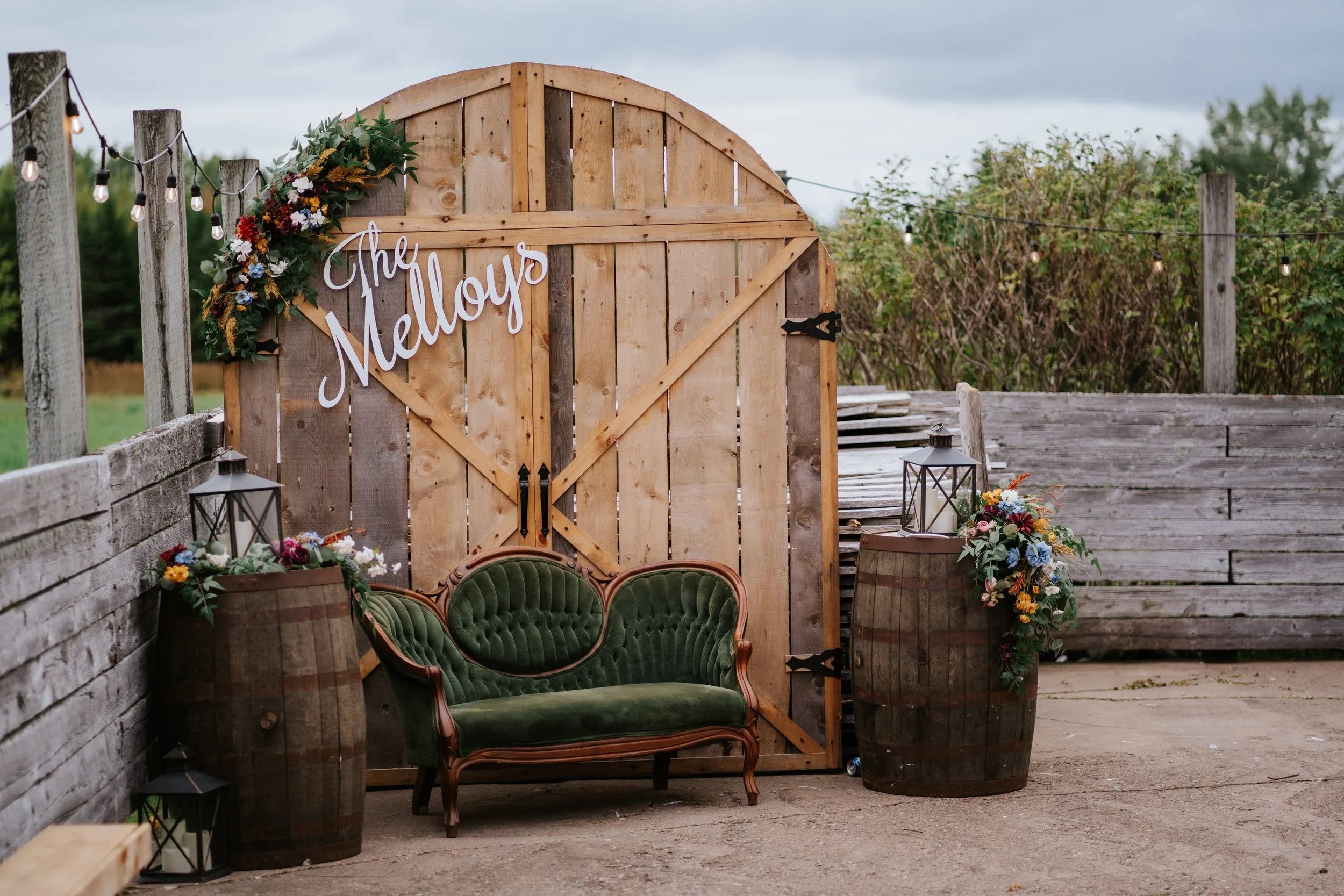 Rustic outdoor wedding decor with a wooden barn door featuring a floral arrangement and the name 'The Mellows,' flanked by vintage barrels, lanterns, and string lights.