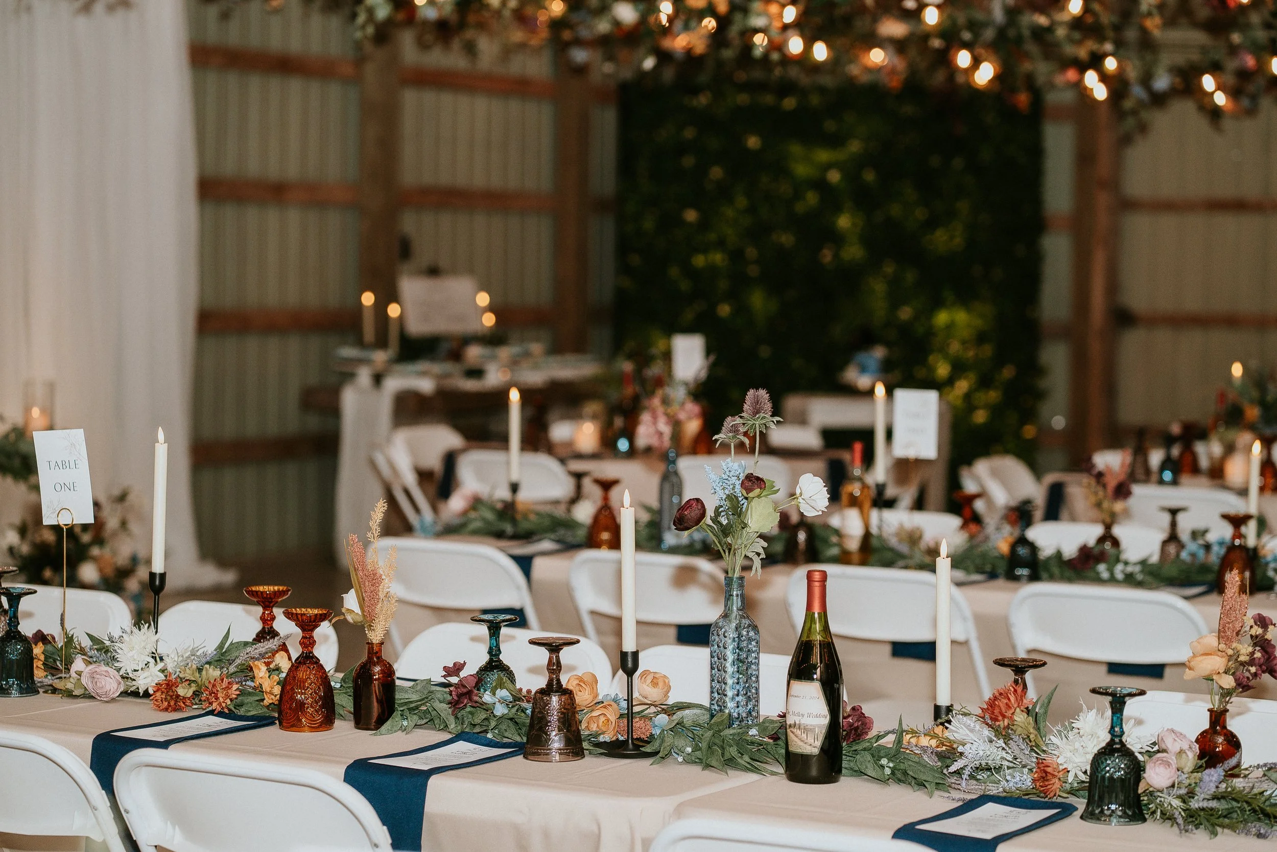 A banquet table decorated with floral centerpieces, candles, and bottles of wine at a wedding reception or celebration event in a rustic barn setting.