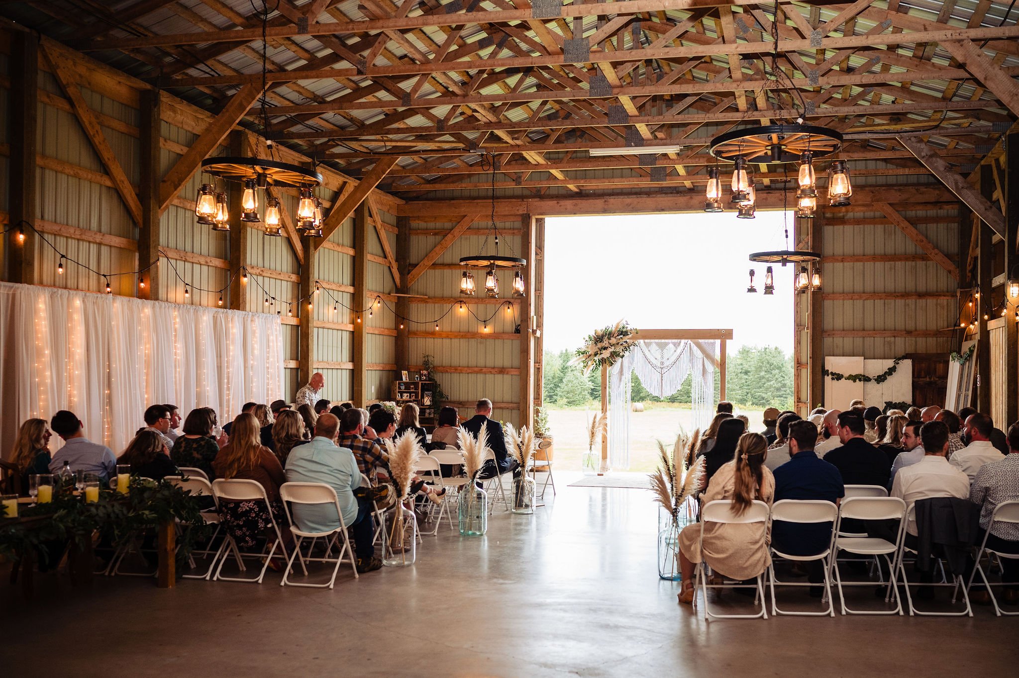 Wedding ceremony setup in a rustic barn with guests seated facing an outdoor scene with a decorative arch.