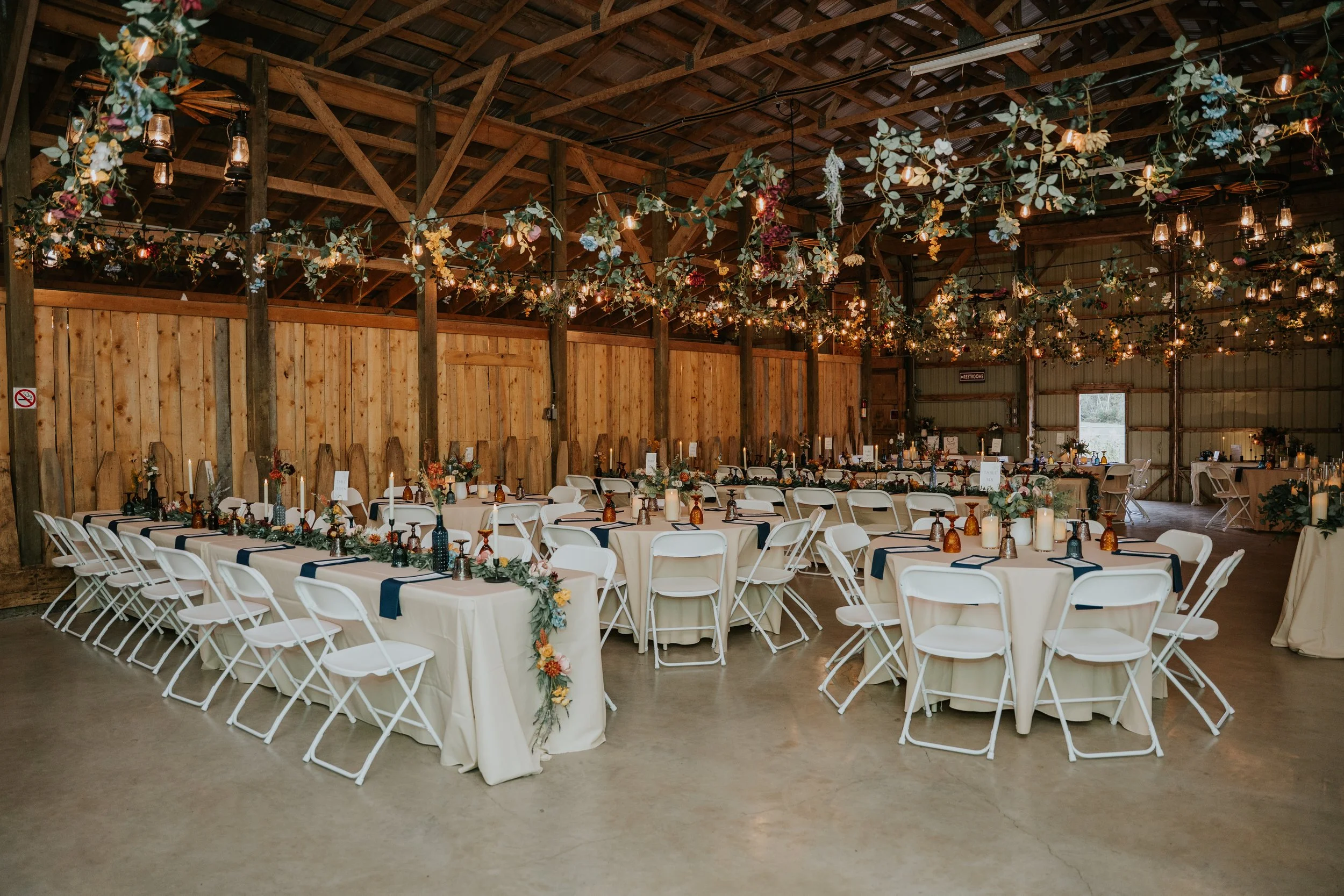 Wedding reception setup in a rustic barn with long and round tables decorated with floral garlands, candles, and small vases, adorned with navy blue napkins, surrounded by white folding chairs. Overhead, strings of small lanterns and hanging floral a