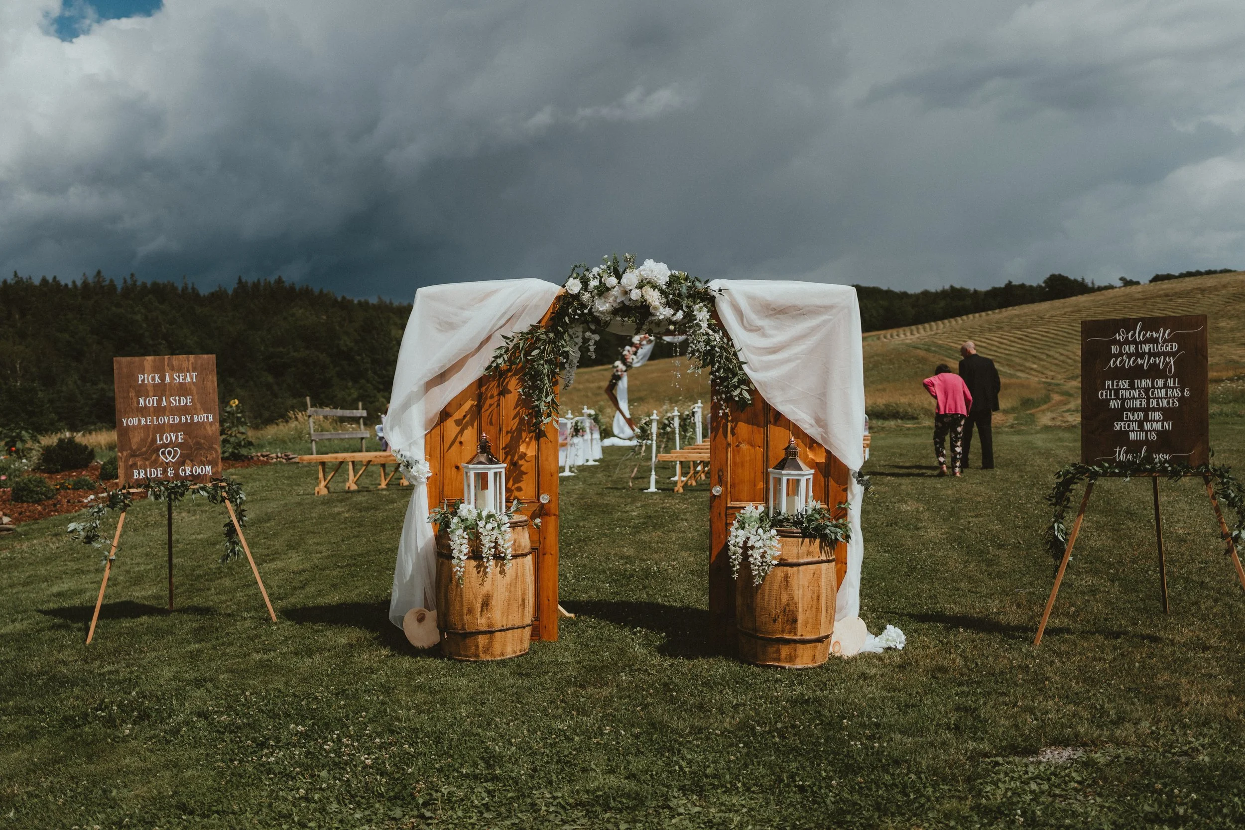 Outdoor wedding ceremony setup on a grassy field with a wooden arch decorated with white drapes and flowers, lanterns, and signs welcoming guests and providing seating instructions.