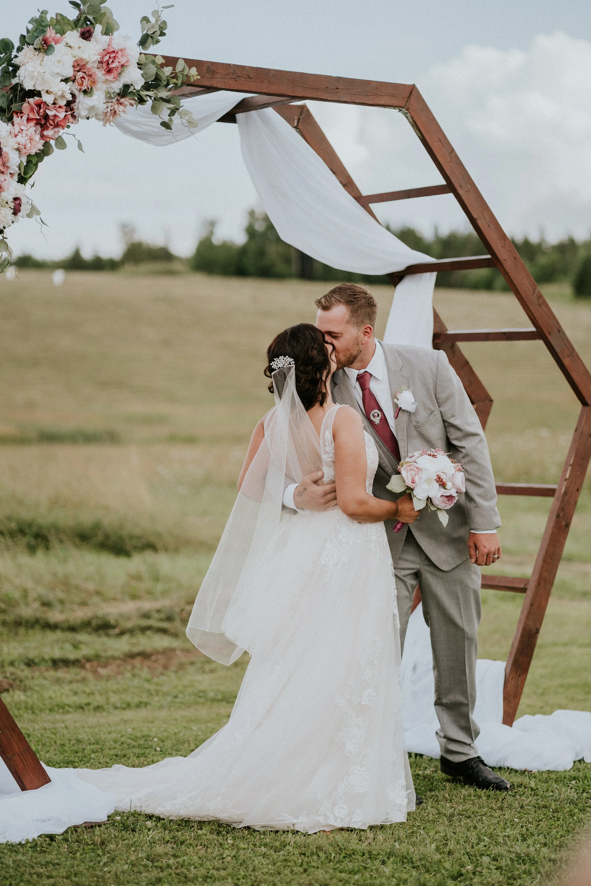 A newlywed couple sharing a kiss during their outdoor wedding ceremony, with a wooden geometric arch decorated with pink and white flowers and white drapery, on a grassy field under a cloudy sky.