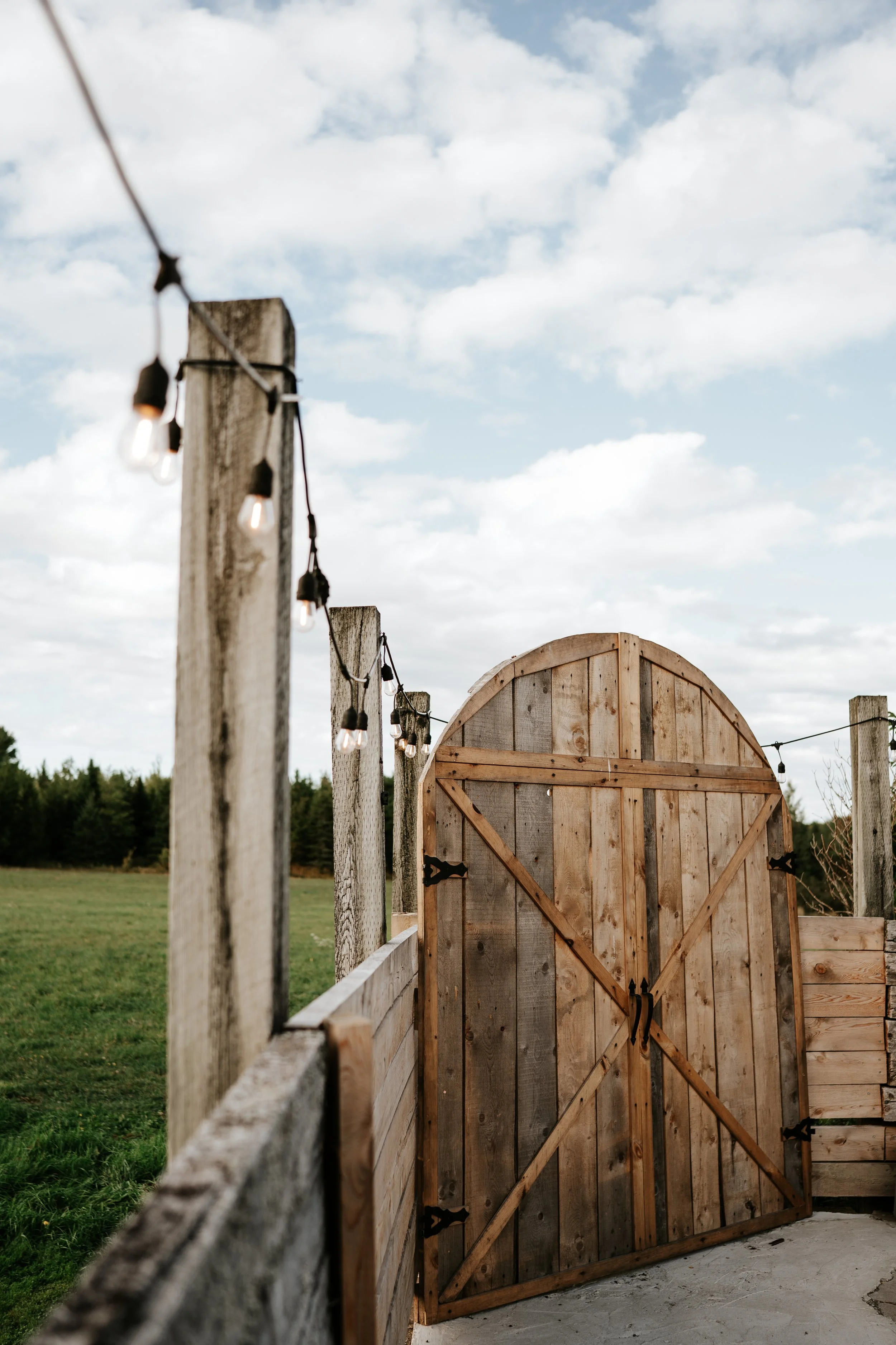 A wooden gate on a farm or rural property with string lights hanging along a weathered wooden fence, open to a grassy field and partly cloudy sky.