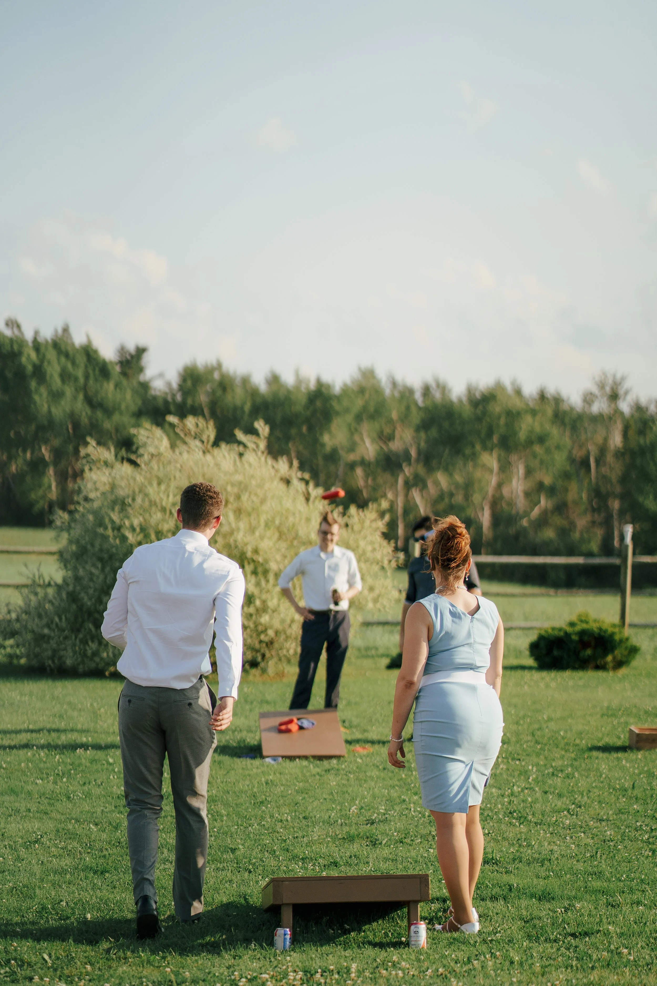 People playing washer toss outdoors on a sunny day, with a woman in a blue dress and two men, one in a white shirt and the other in a dark shirt, participating in the game.