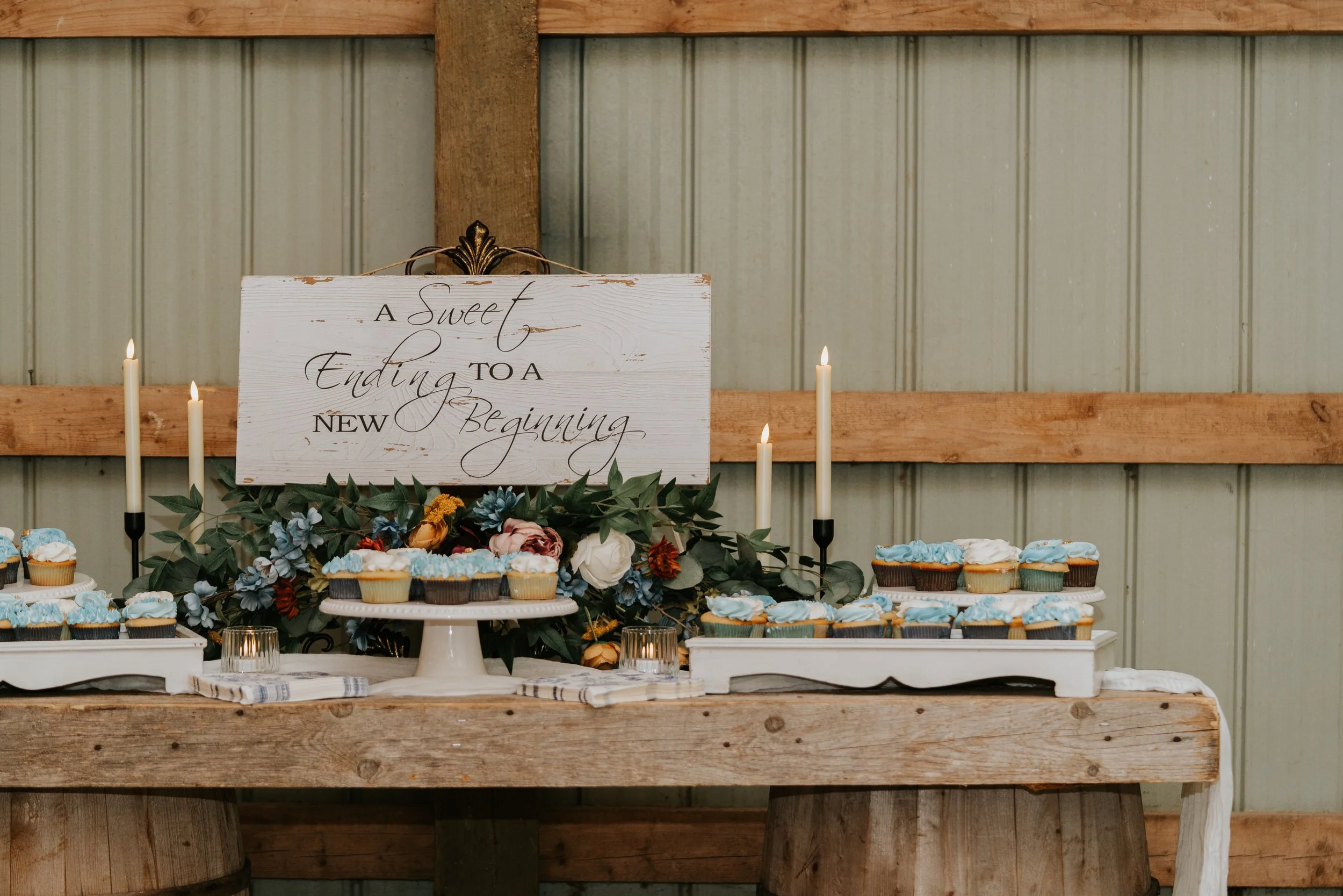 Table decorated with cupcakes, candles, and flowers against a rustic wooden backdrop, featuring a sign with the message 'A sweet ending to a new beginning'.