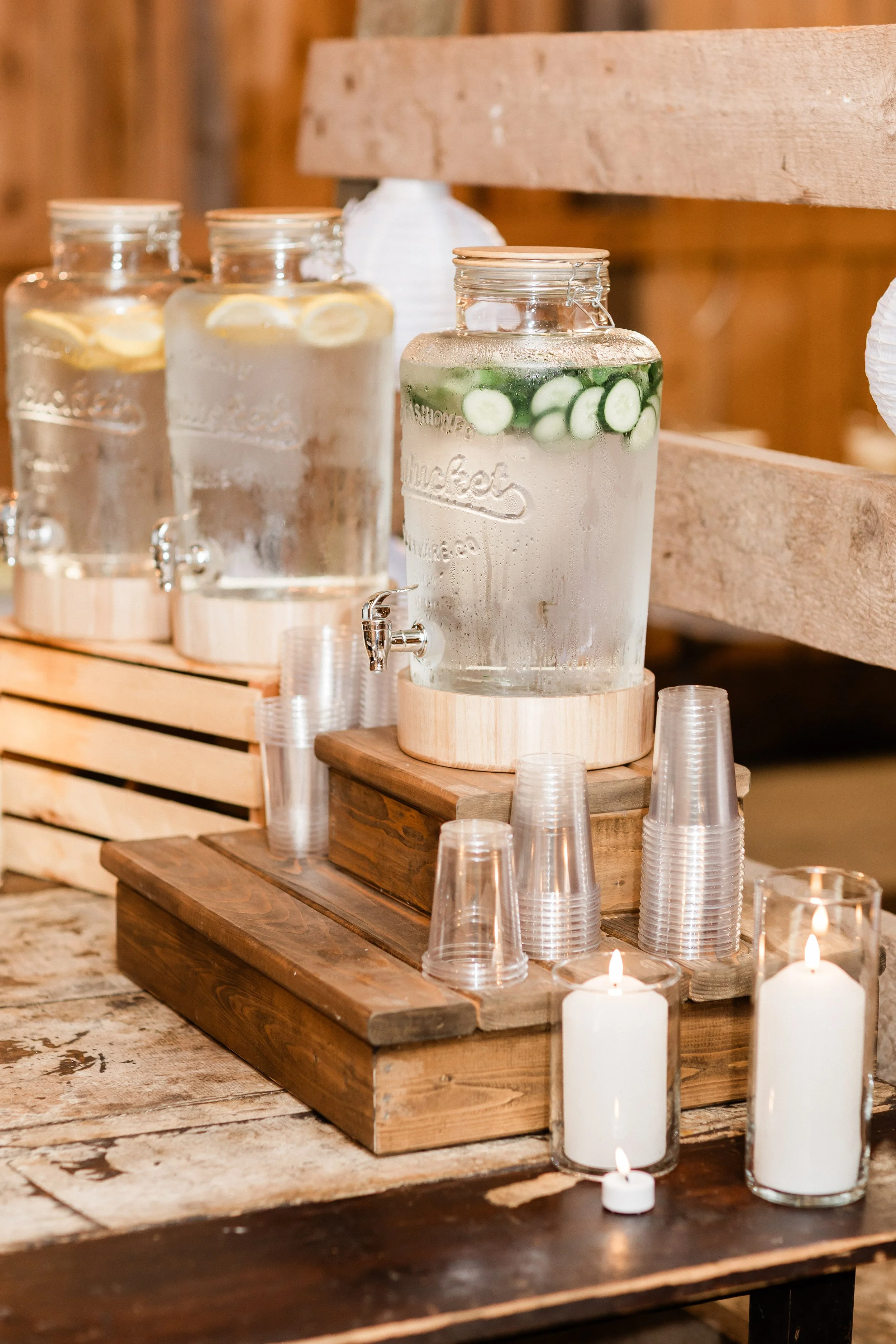 Three large glass jars of infused water with lemon and cucumber slices, placed on wooden slabs, surrounded by disposable cups and candles on a rustic wooden table.