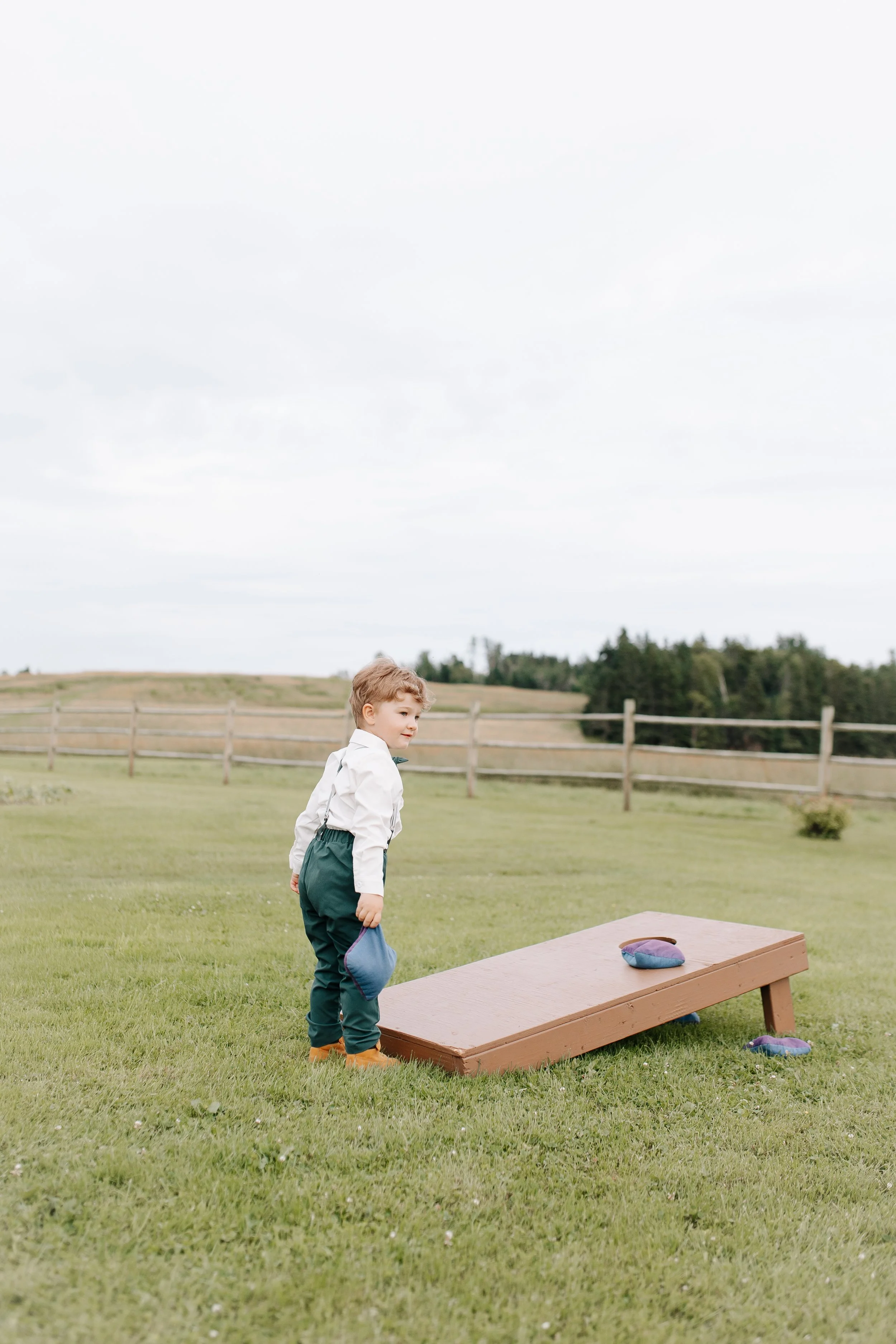 Young boy in dress shirt, pants, and suspenders standing outdoors near a horse-shoe pitching game on grass, with wooden fence and rolling hills in the background.