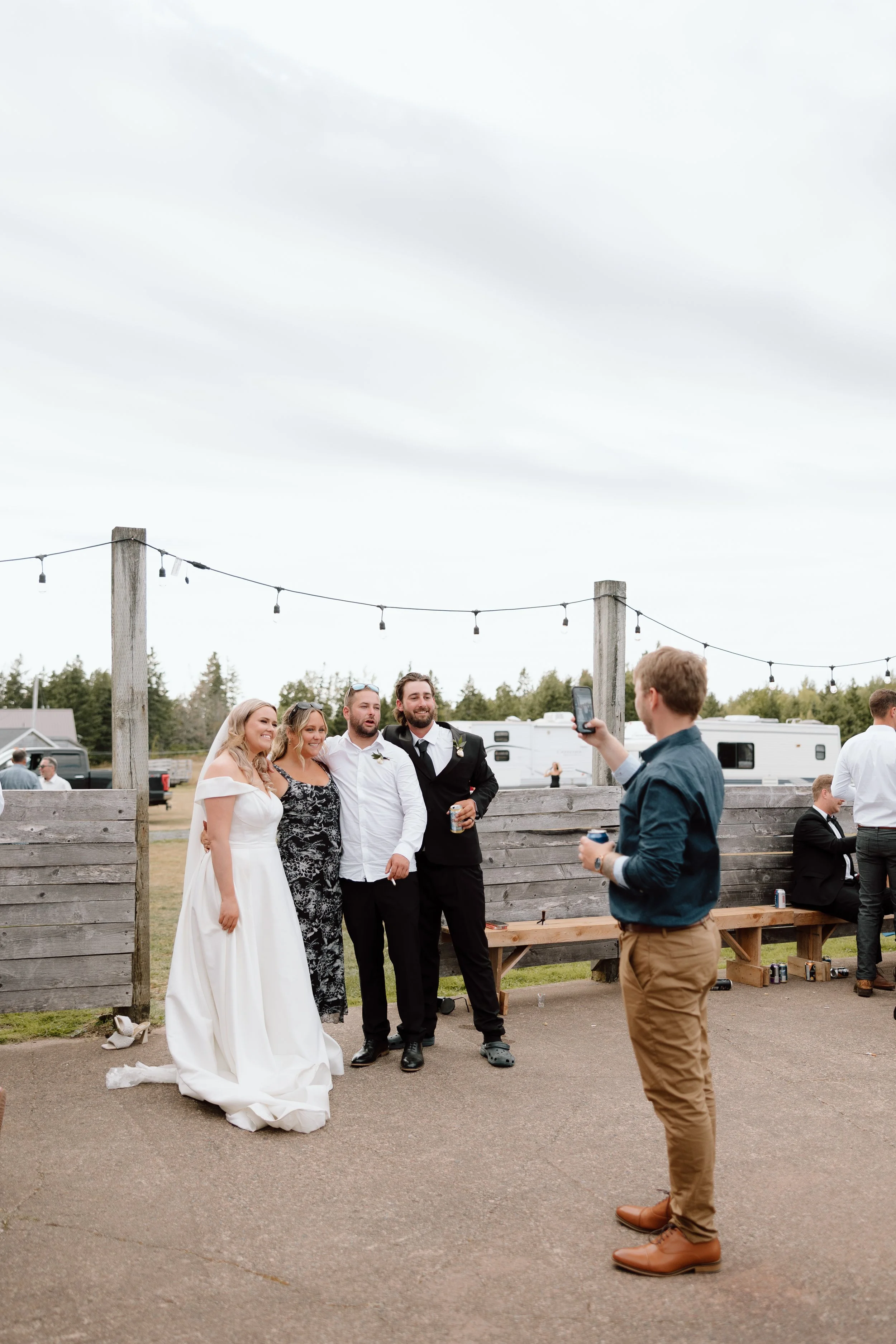 Wedding guests taking a photo of a bride and groom at an outdoor reception with string lights and a wooden fence.