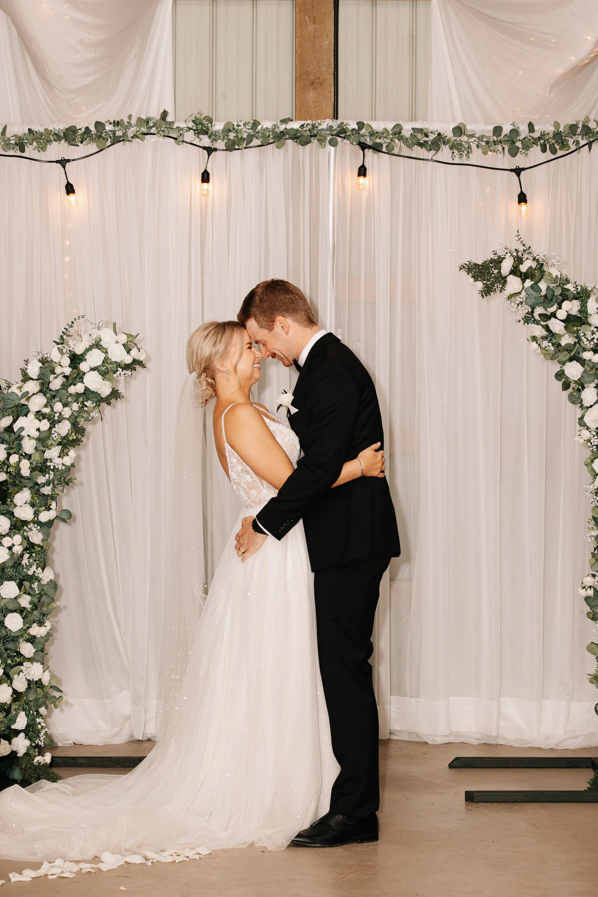 A bride and groom share a kiss at their wedding ceremony, standing under a decorated arch with white flowers and greenery, with string lights overhead.
