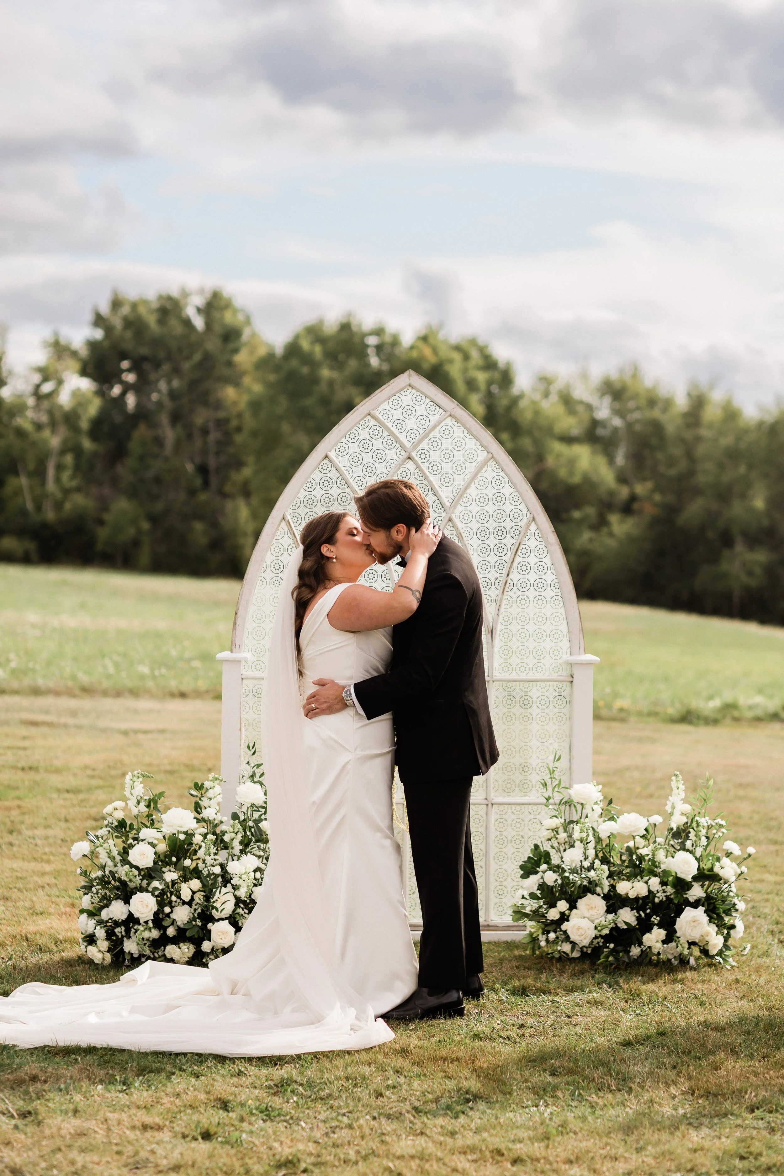 A bride and groom kiss during an outdoor wedding ceremony in a field with trees in the background, under a partly cloudy sky, with floral arrangements and a decorative backdrop.