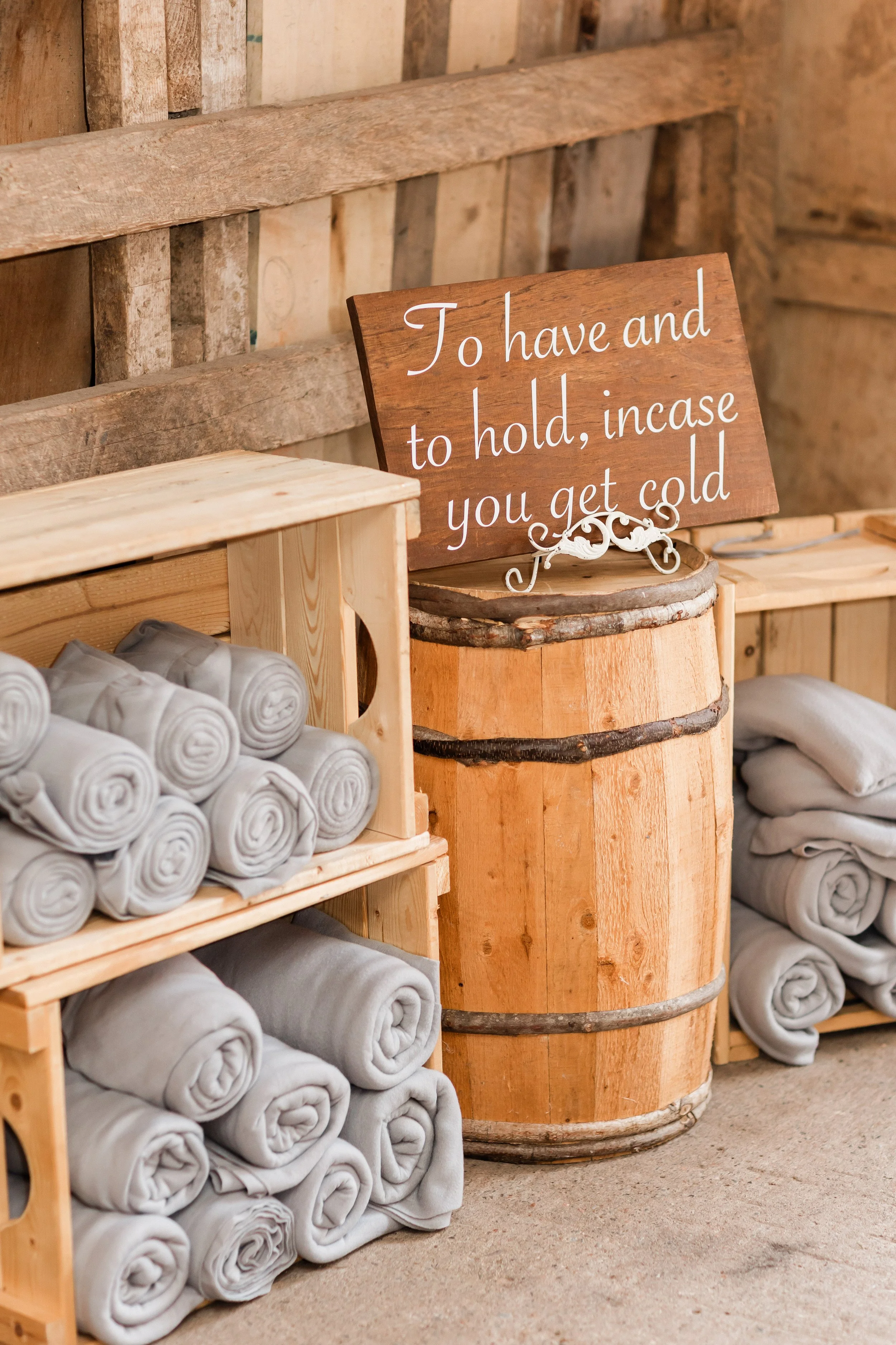 Stacked white rolled towels and folded blankets on wooden shelves, with a wooden barrel and a sign that reads 'To have and to hold, incase you get cold' in a rustic setting.