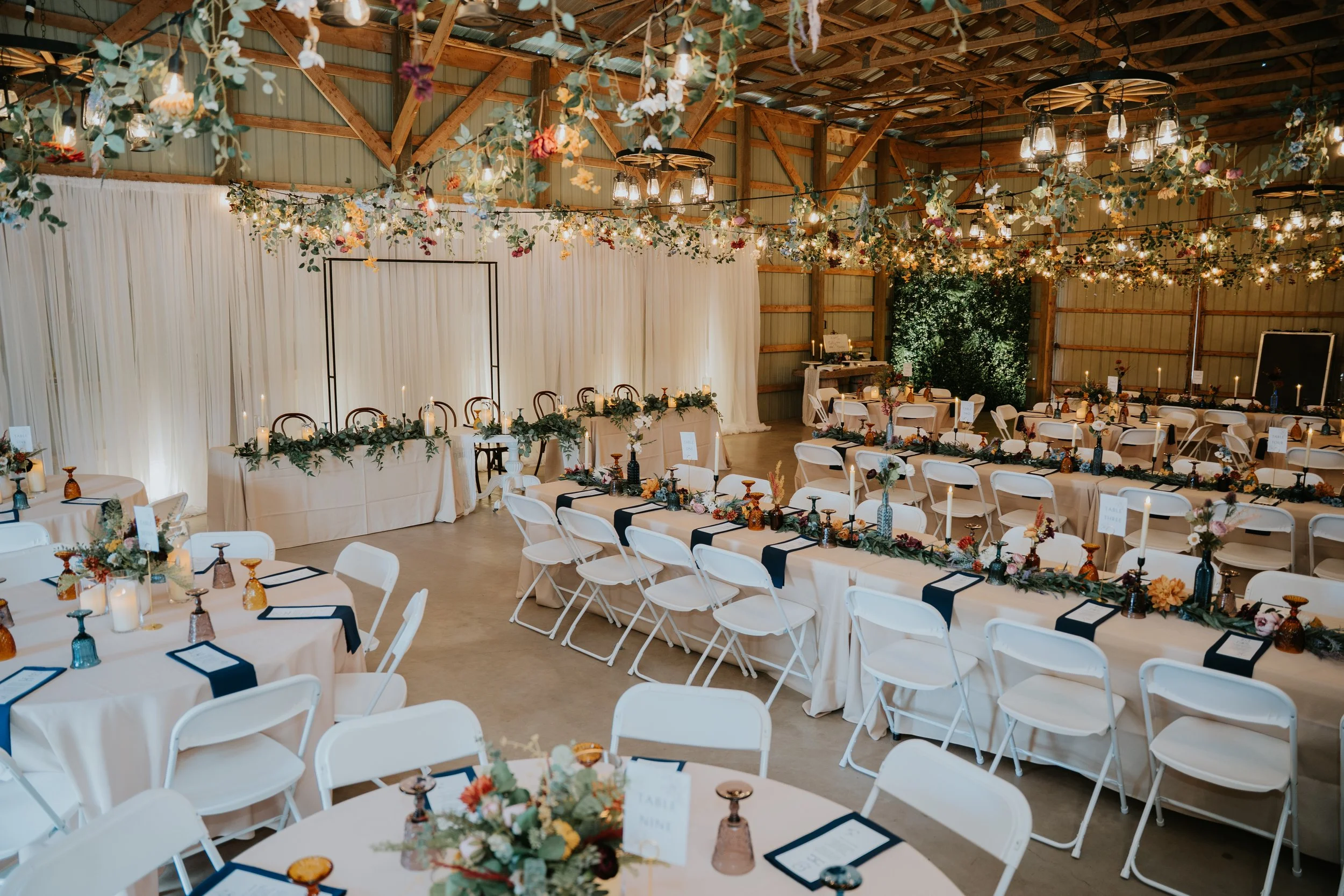 Wedding reception setup in a barn with decorated tables, floral arrangements, candleholders, and hanging floral and greenery decor with string lights.