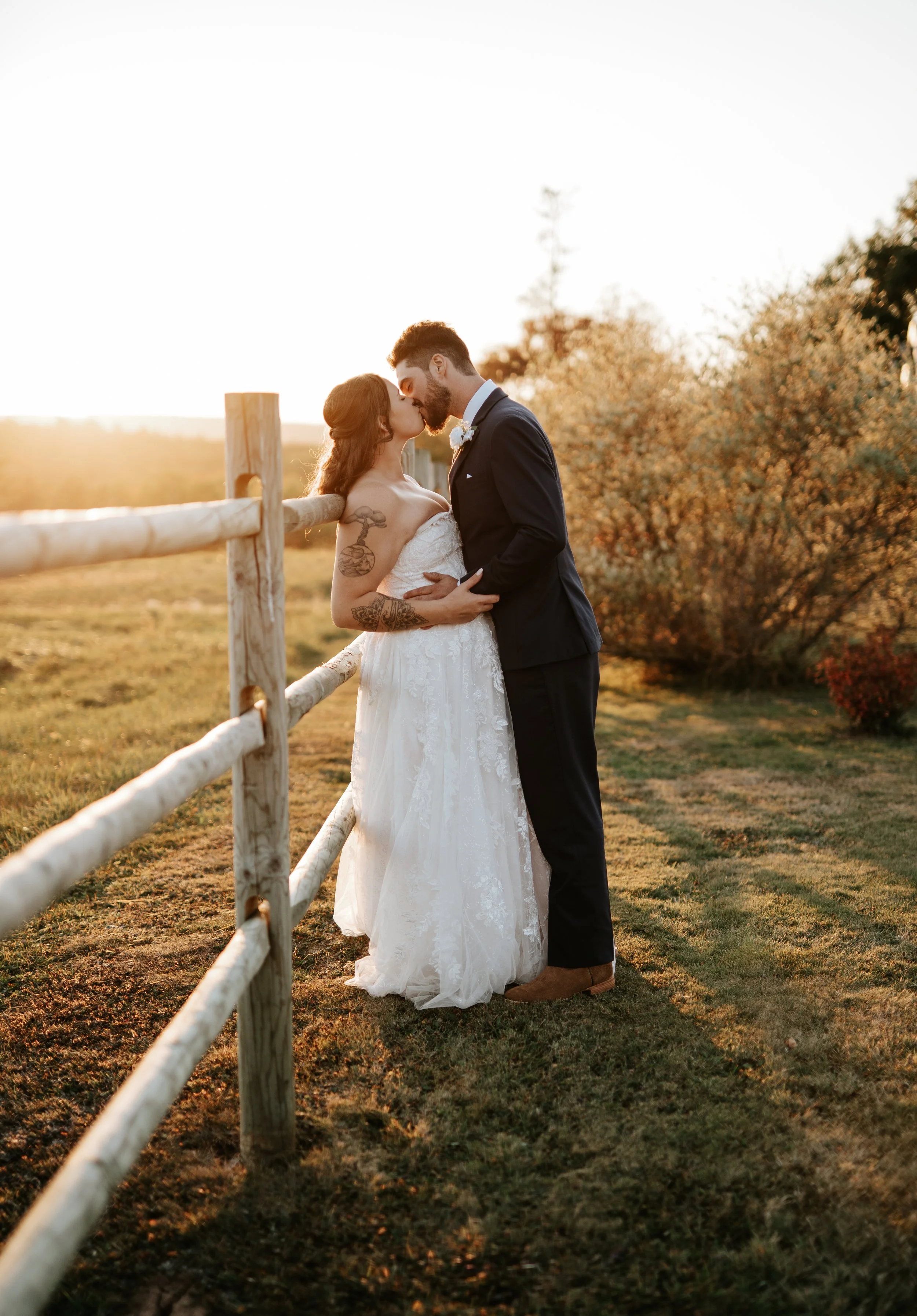 A newlywed couple sharing a kiss outdoors at sunset, with a wooden fence and trees in the background.