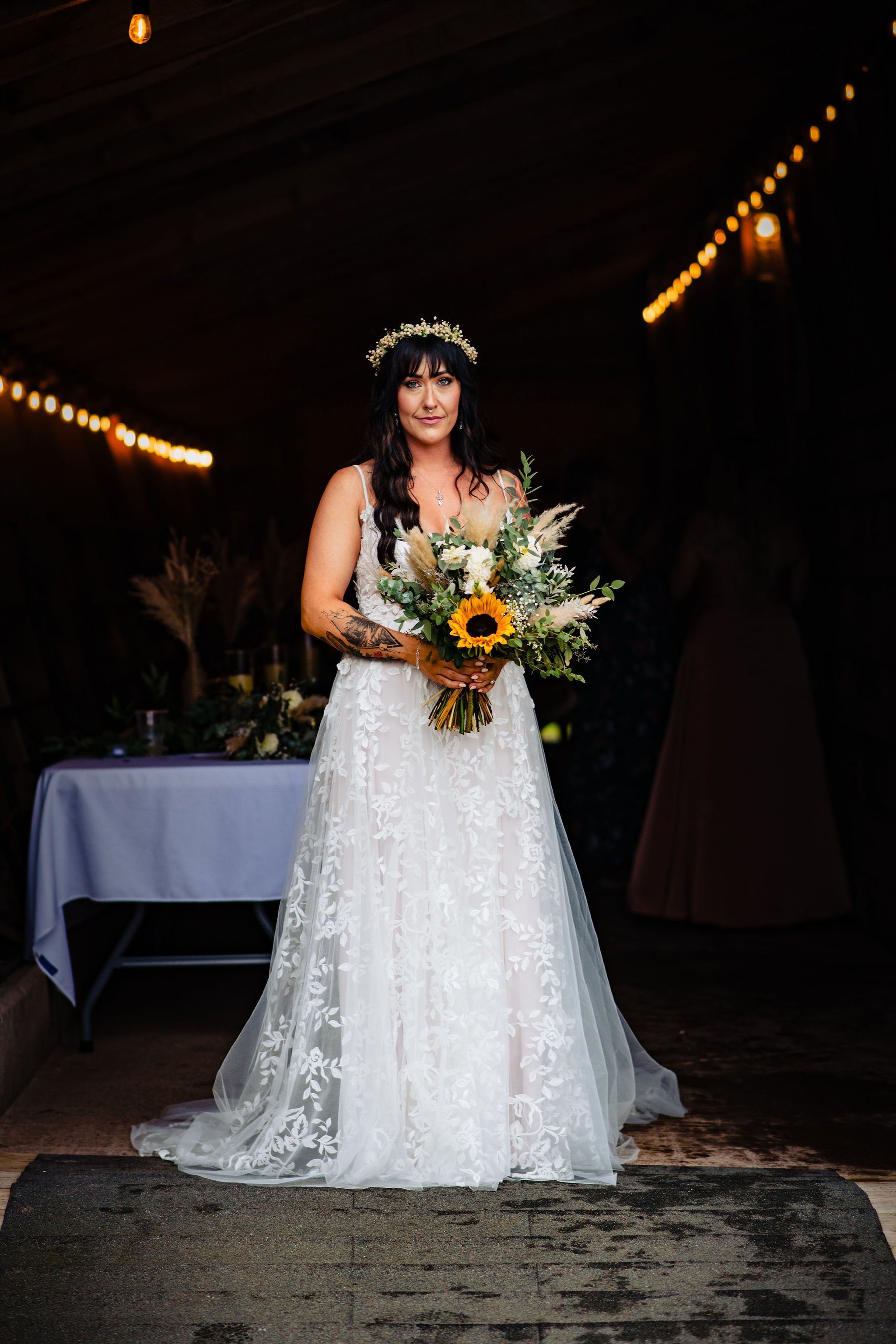 A bride in a white lace wedding dress holding a bouquet of sunflowers and greenery, standing indoors with string lights overhead.