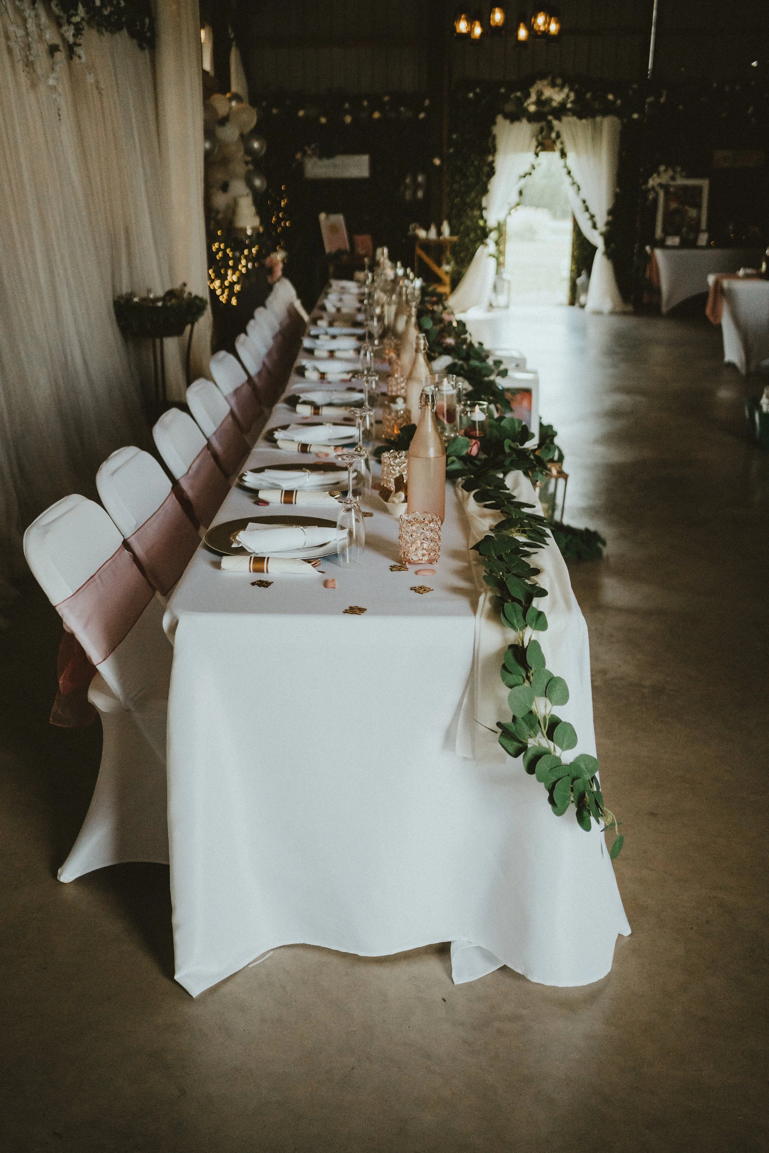A long rectangular table set for a wedding reception with white tablecloth, white chairs with pink sashes, and decor including pink bottles, candles, and green foliage, inside a decorated venue with curtains and fairy lights.