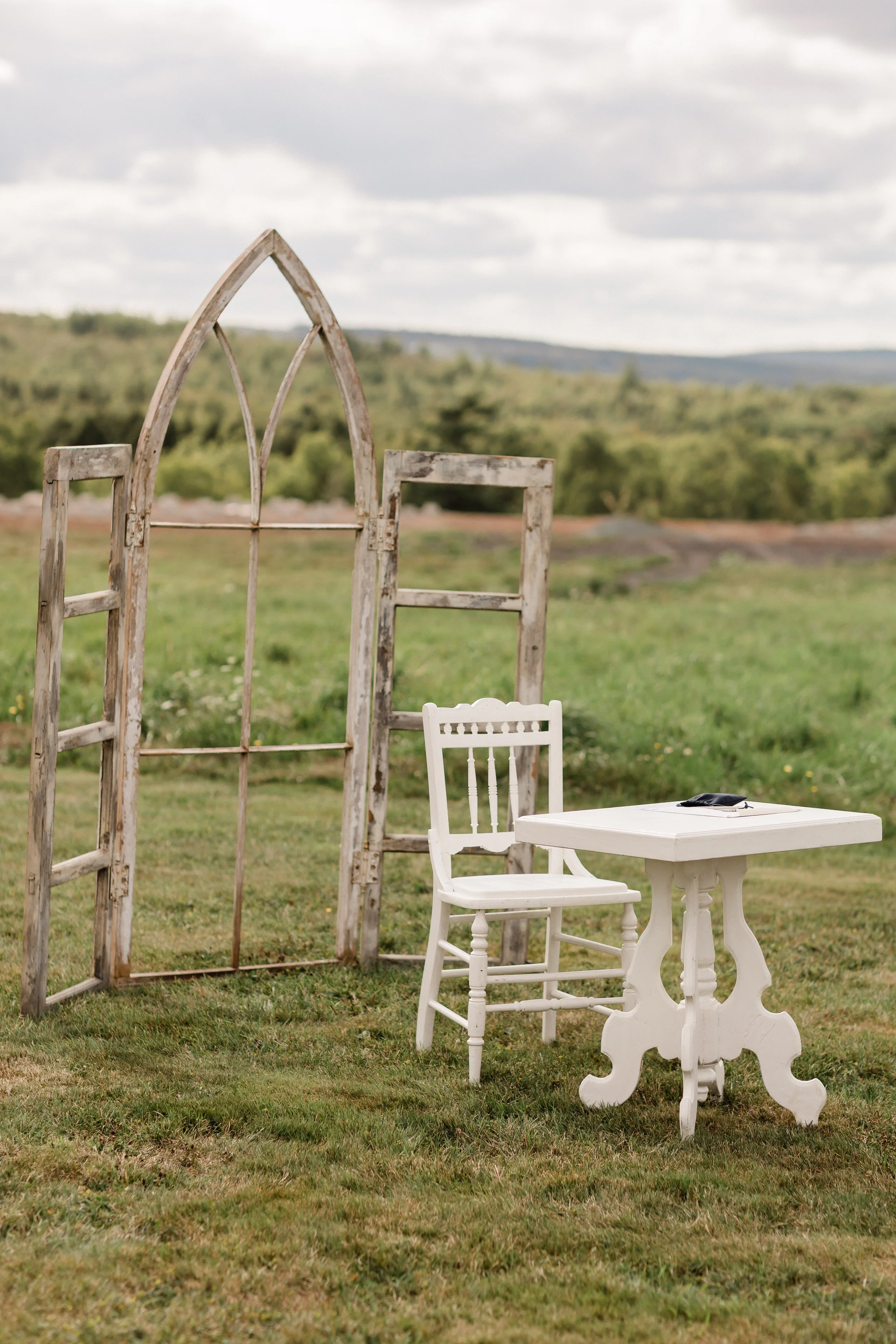 An outdoor scene with a white carved wooden table, a white chair, a rustic wooden ladder, and a window frame set on grass, with a backdrop of green fields and a cloudy sky.