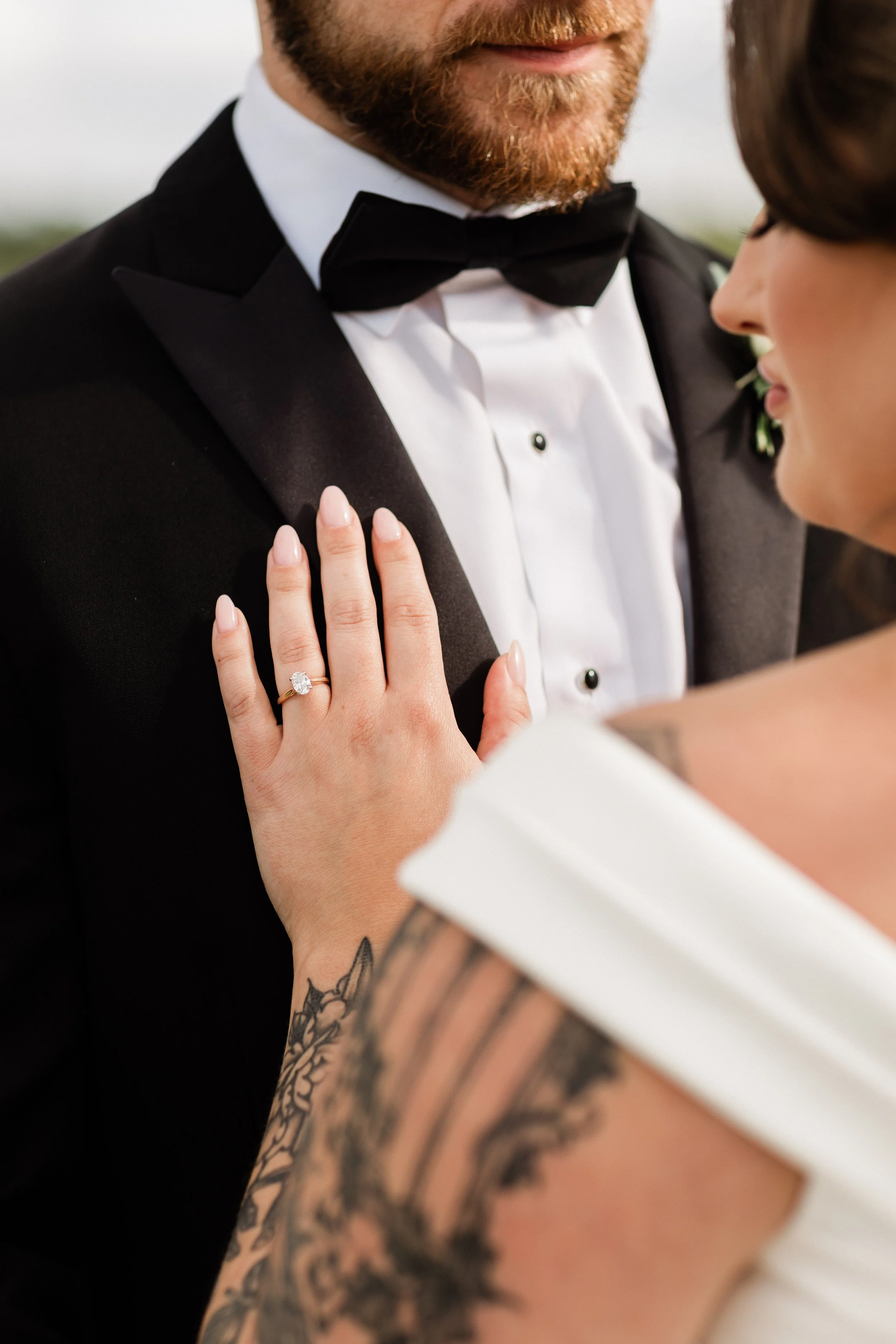 Close-up of a newlywed couple, with the bride's hand on the groom's chest, showcasing an engagement ring. The groom is wearing a black tuxedo with a black bow tie, and the bride has a tattooed arm visible.