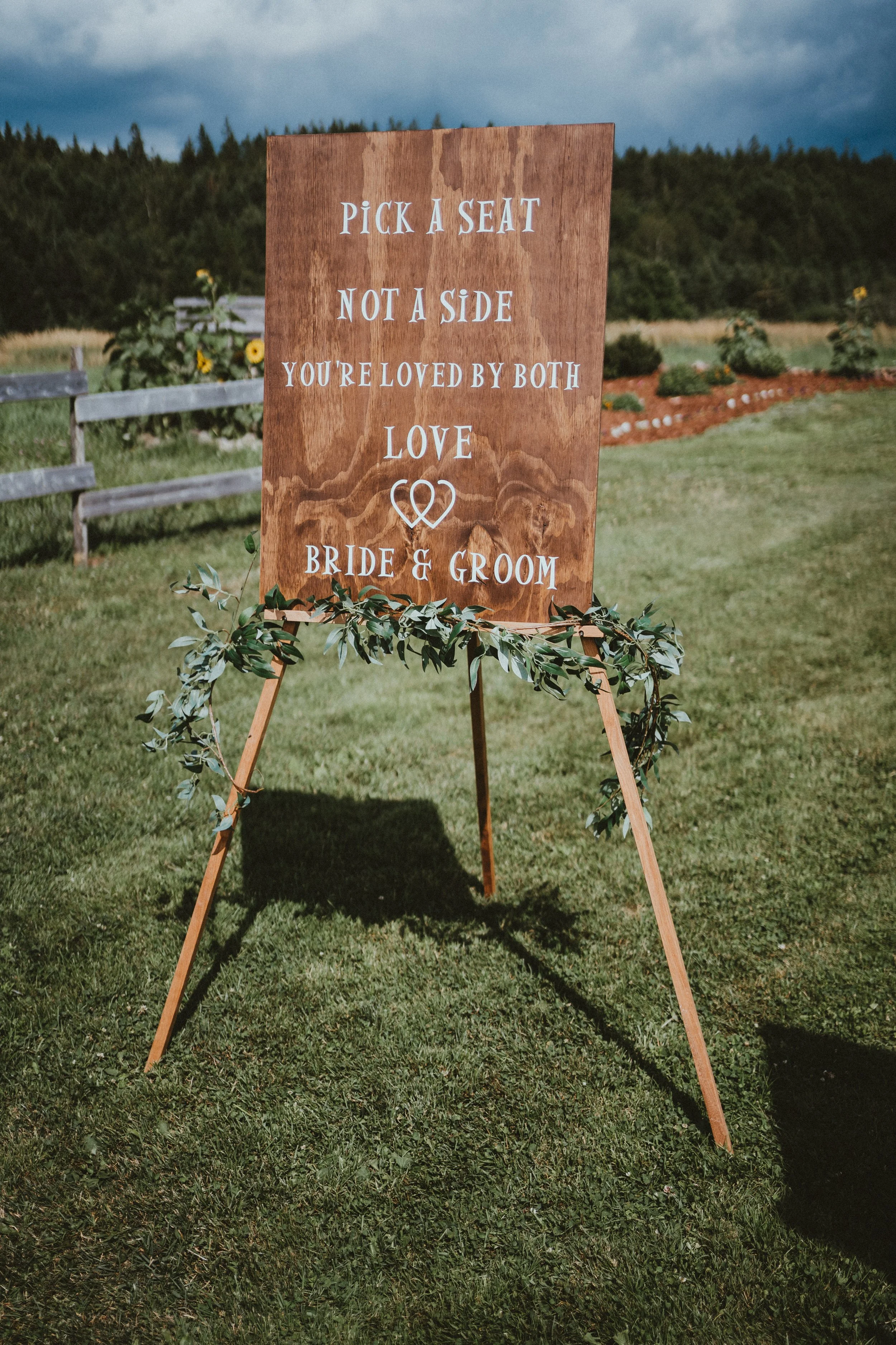 A wooden wedding sign on an easel with greenery, standing on a grassy field with trees and a fence in the background. The sign reads: 'Pick a seat not a side. You're loved by both. Love, Bride & Groom.'