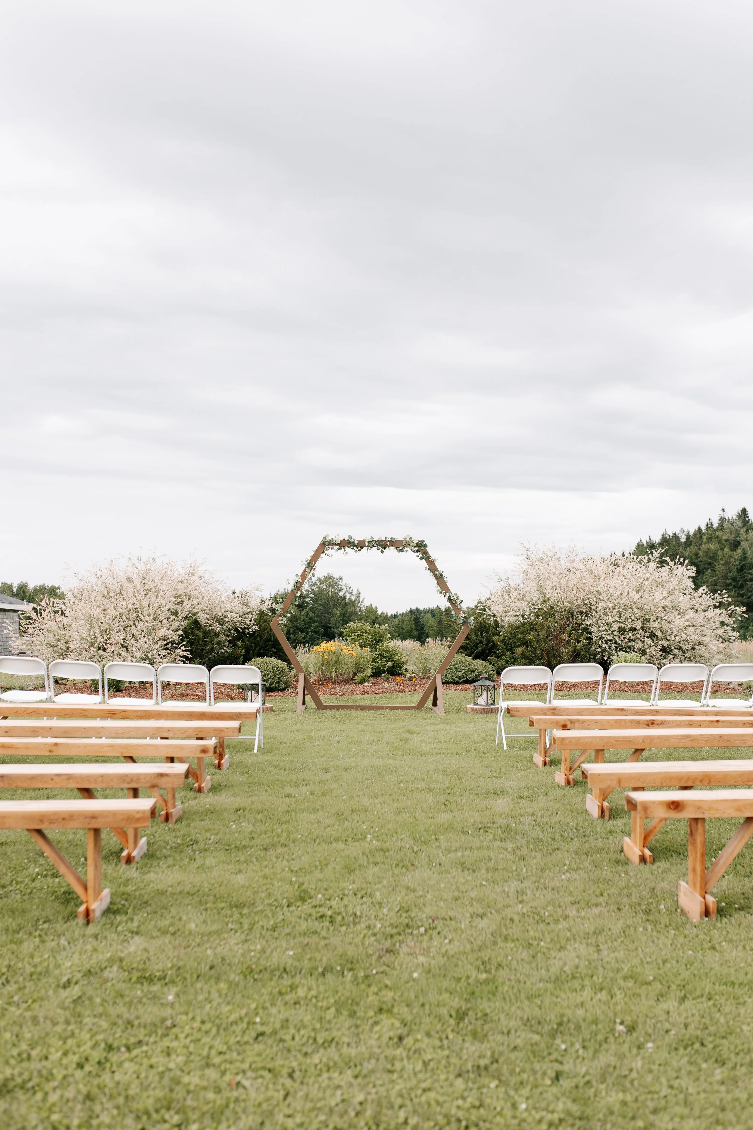 Outdoor wedding ceremony setup with wooden benches and white folding chairs facing a hexagonal wooden arch decorated with greenery, situated on a lush green lawn with blossoming trees in the background under a cloudy sky.