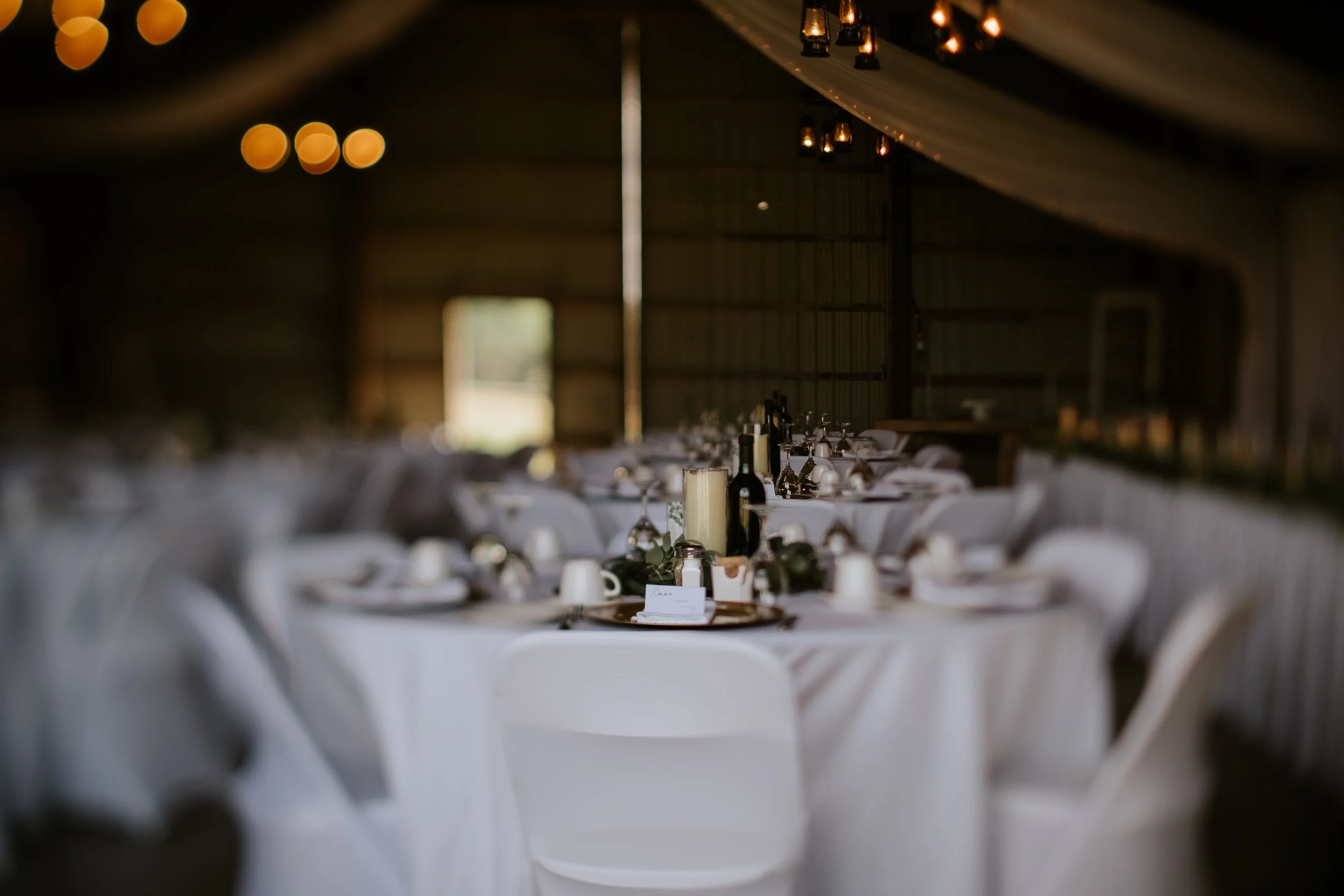 Wedding reception setup inside a barn with round tables covered in white tablecloths, decorated with bottles of wine, candles, and place settings, with warm lighting and a rustic ambiance.