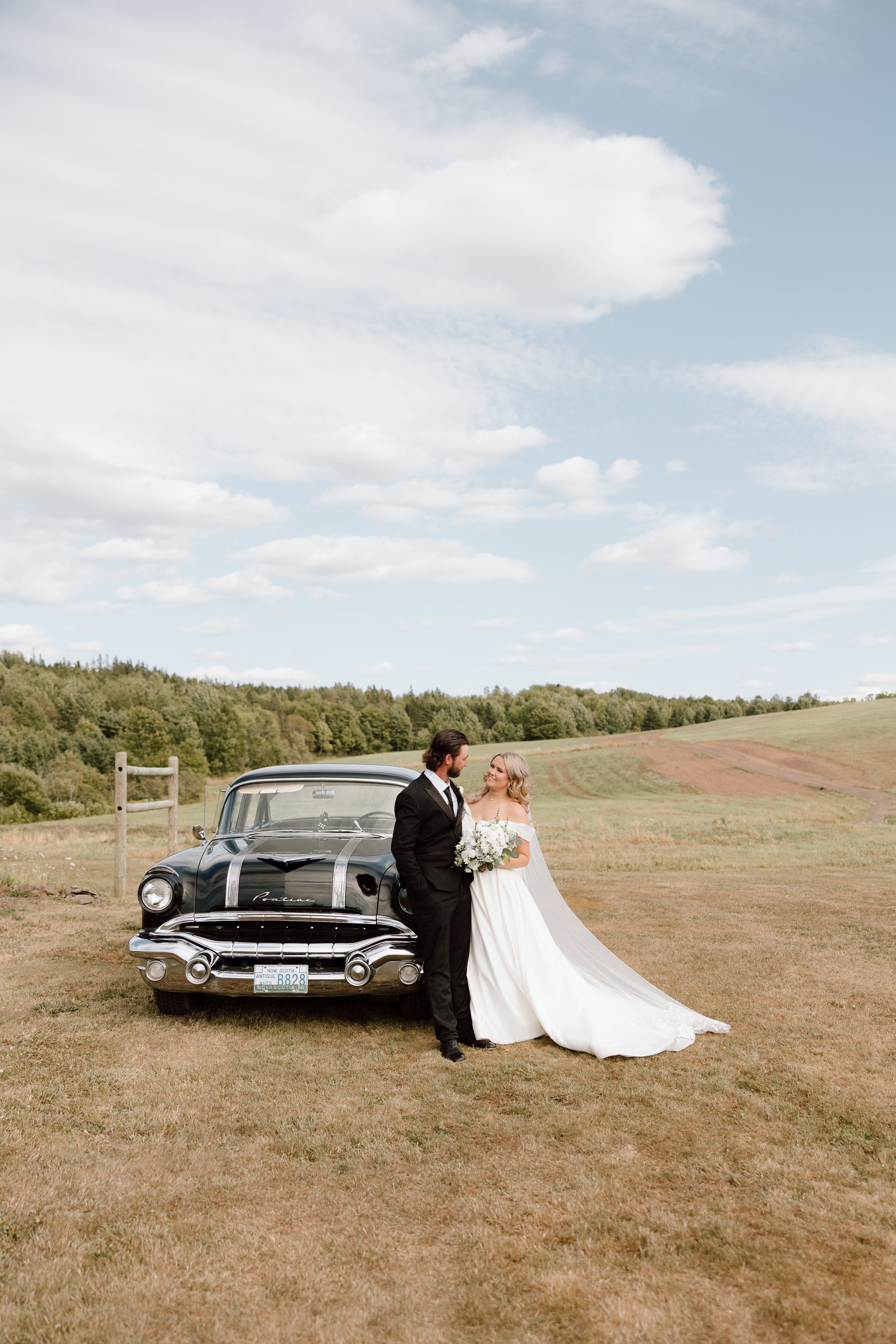Bride and groom standing together outdoors next to a vintage black car on a grassy field with a cloudy sky.