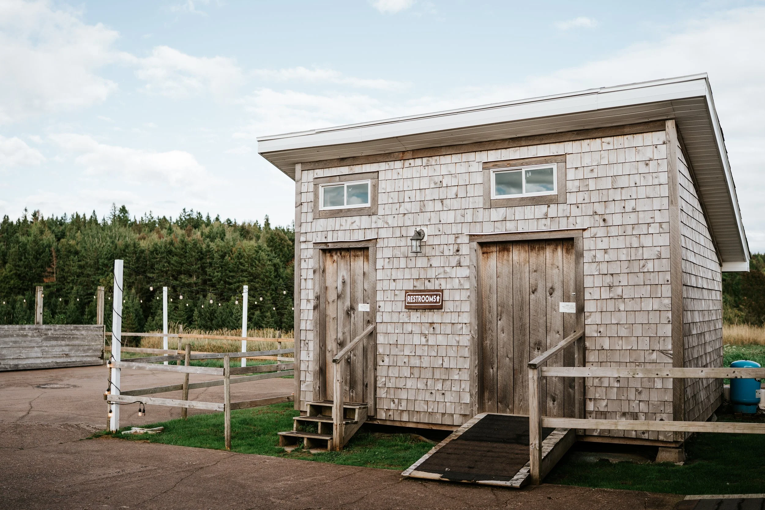 Wooden public restroom building with a ramp and stairs, situated outdoors with surrounding grass and trees in the background.