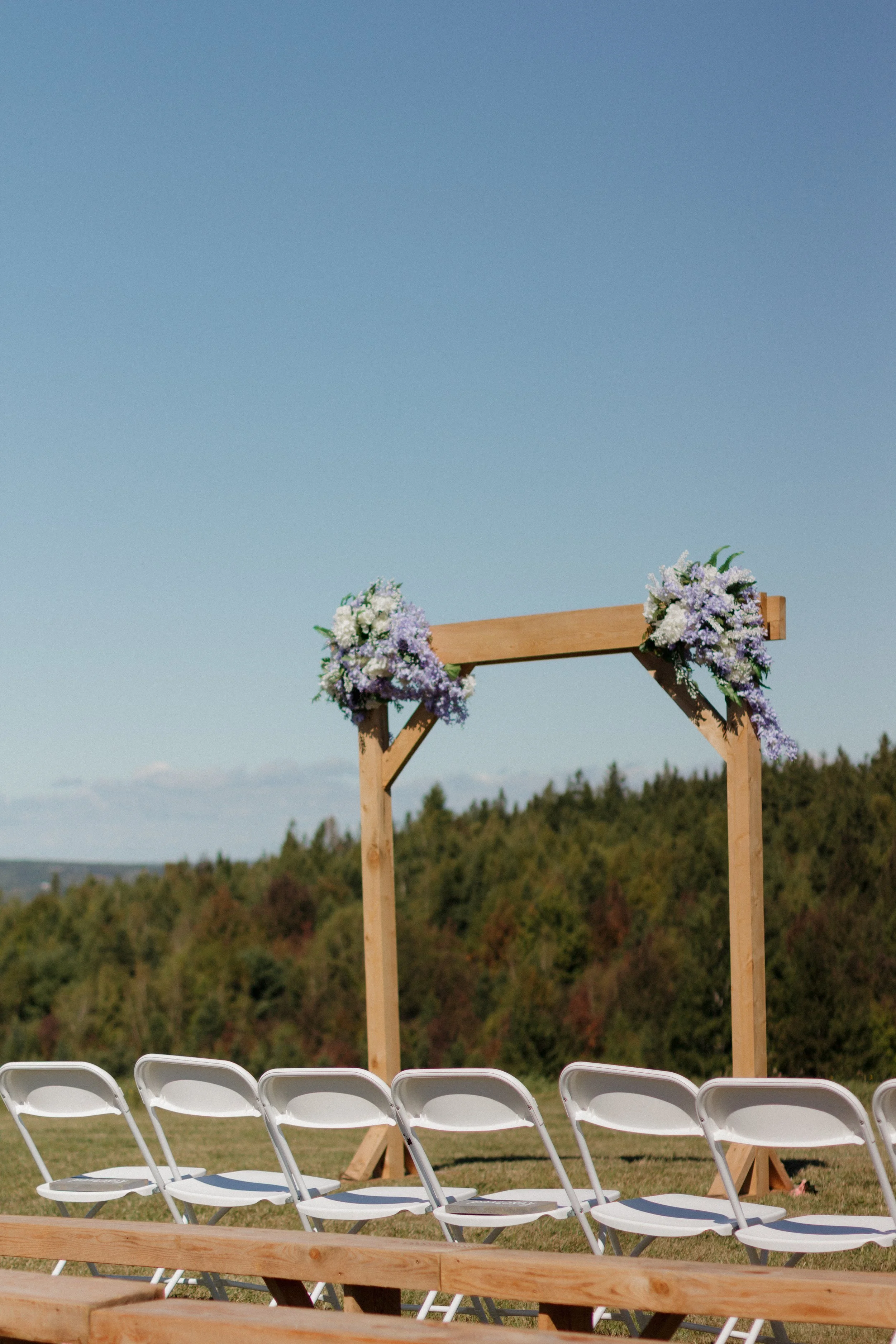 Outdoor wedding ceremony setup with a wooden arbor decorated with purple and white flowers, and white folding chairs arranged on grass under a clear blue sky.