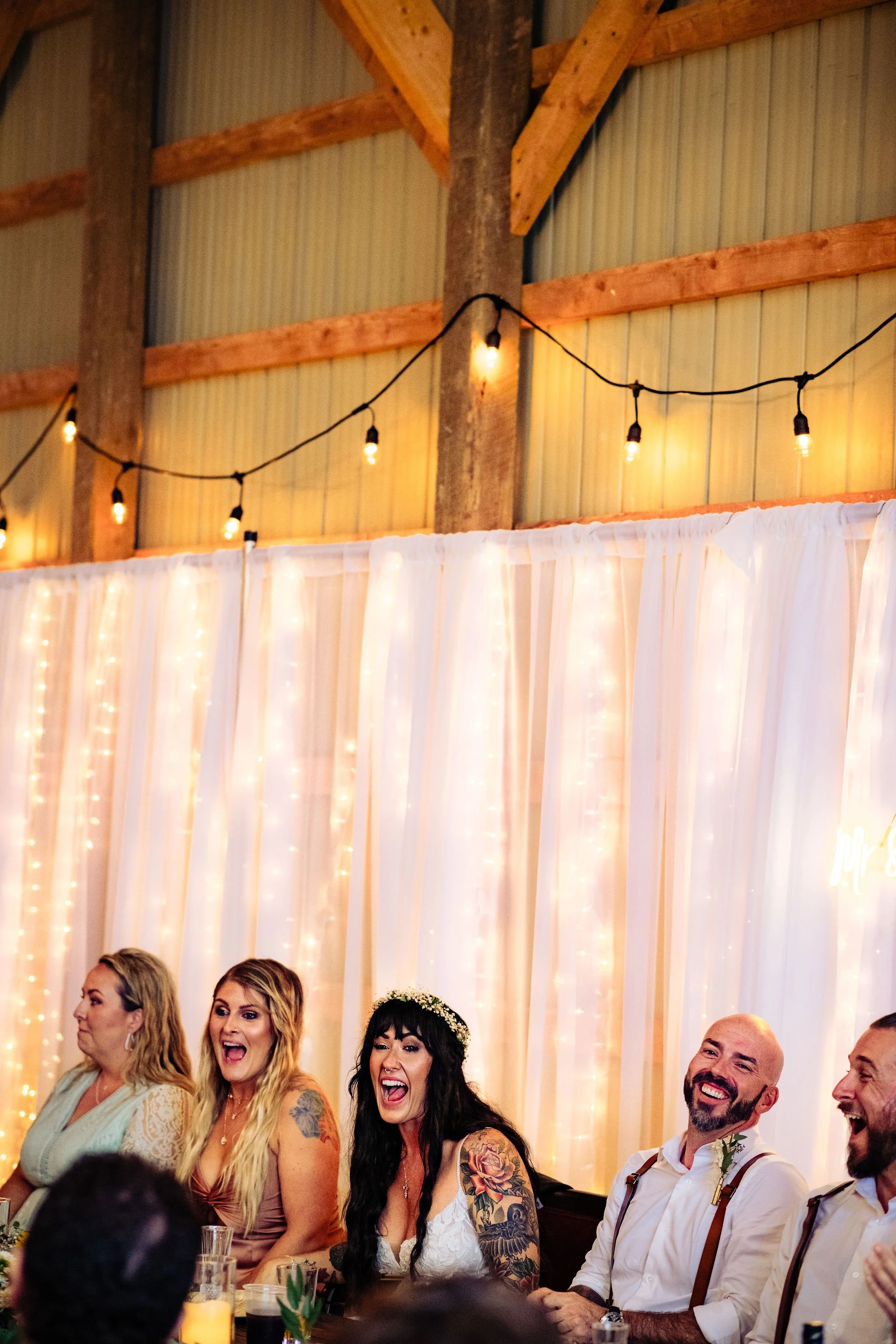 Wedding reception with smiling guests seated at a table, decorated with white drapes and string lights, in a rustic barn setting.