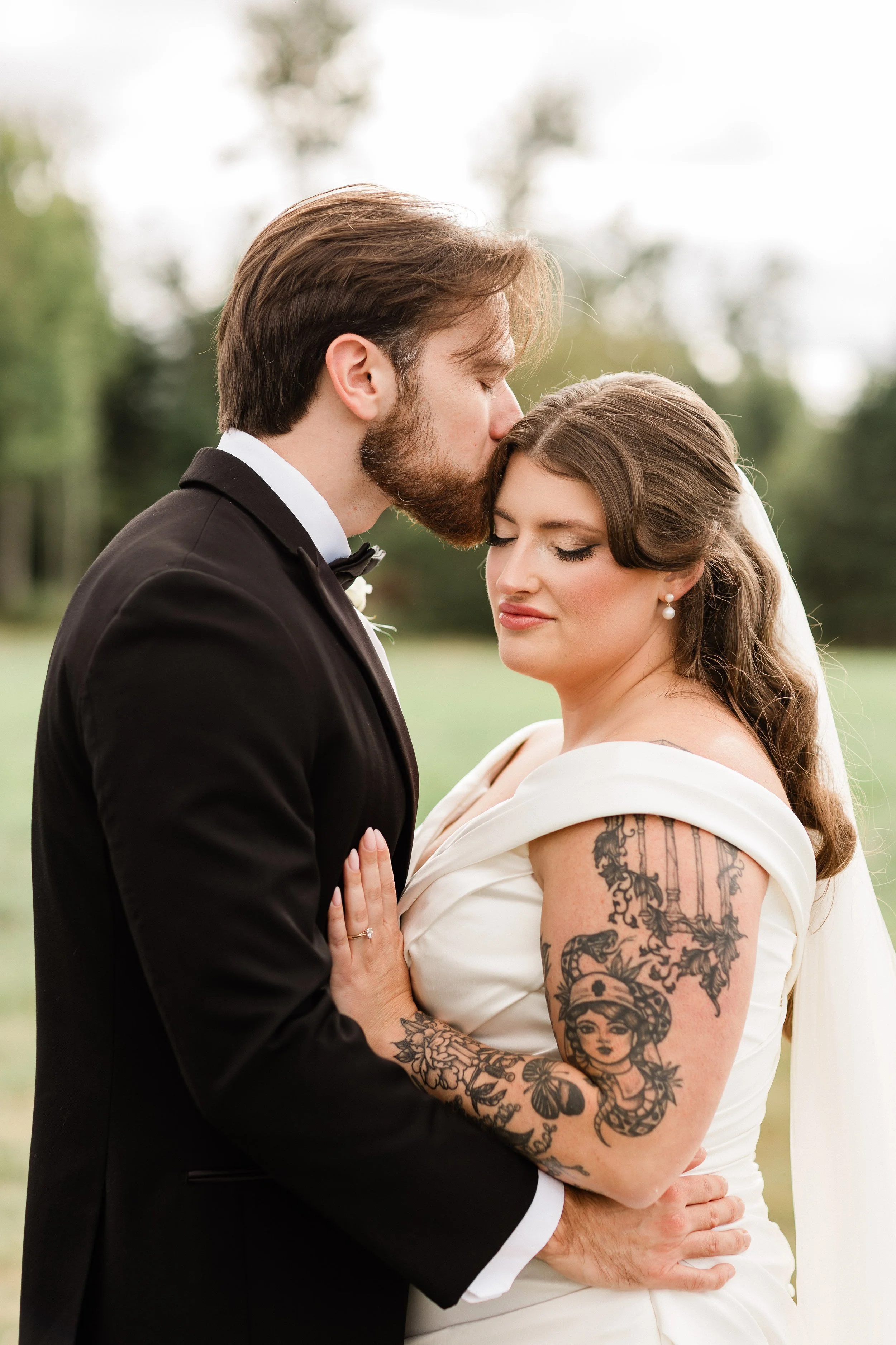 A bride and groom are embracing outdoors. The groom is kissing the bride's forehead, and the bride has her eyes closed. She has tattoos on her arm and is wearing a white wedding dress. The groom is wearing a black tuxedo. The background is blurred gr