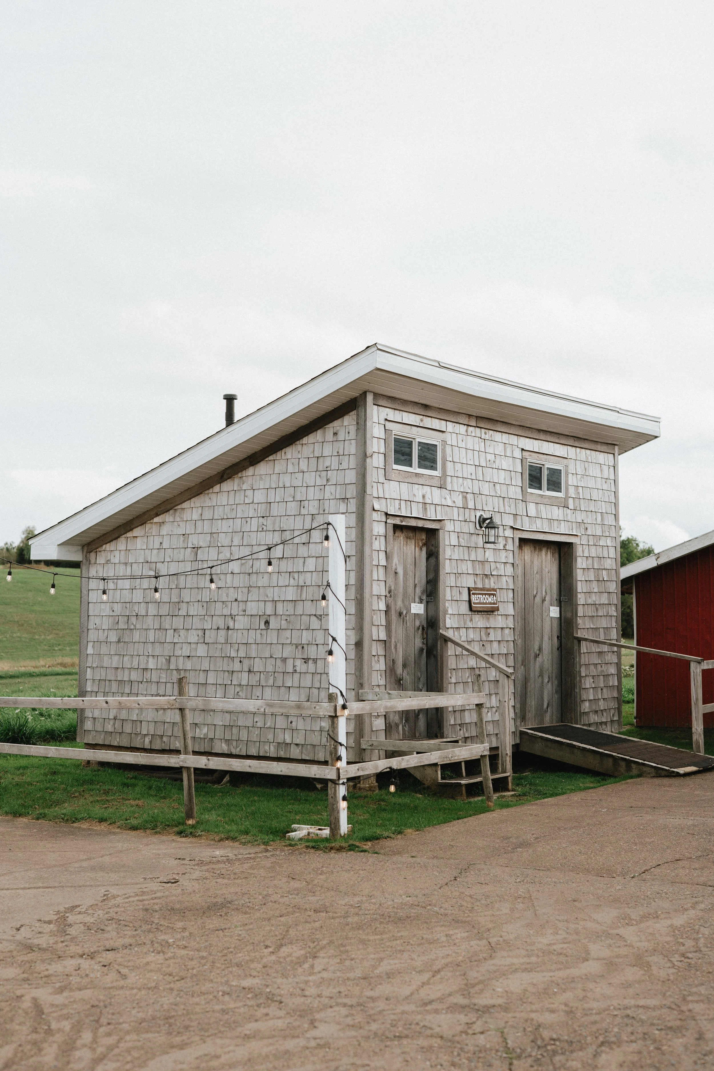 Small rustic building with wooden shingle siding, two upper small windows, two taller side-by-side doors with ramps, string lights hanging outside, situated on a concrete and grassy area.