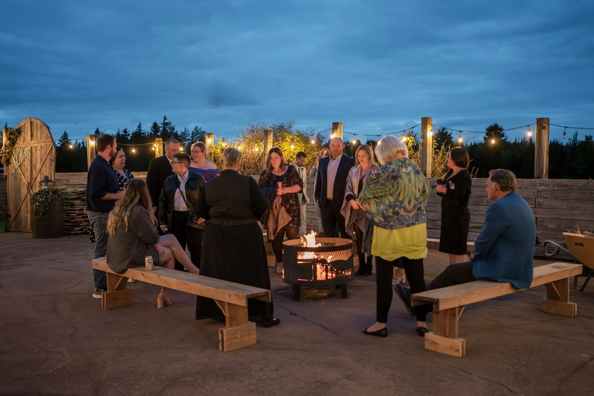 Group of people around a fire pit at an outdoor evening gathering with string lights and a wooden fence.