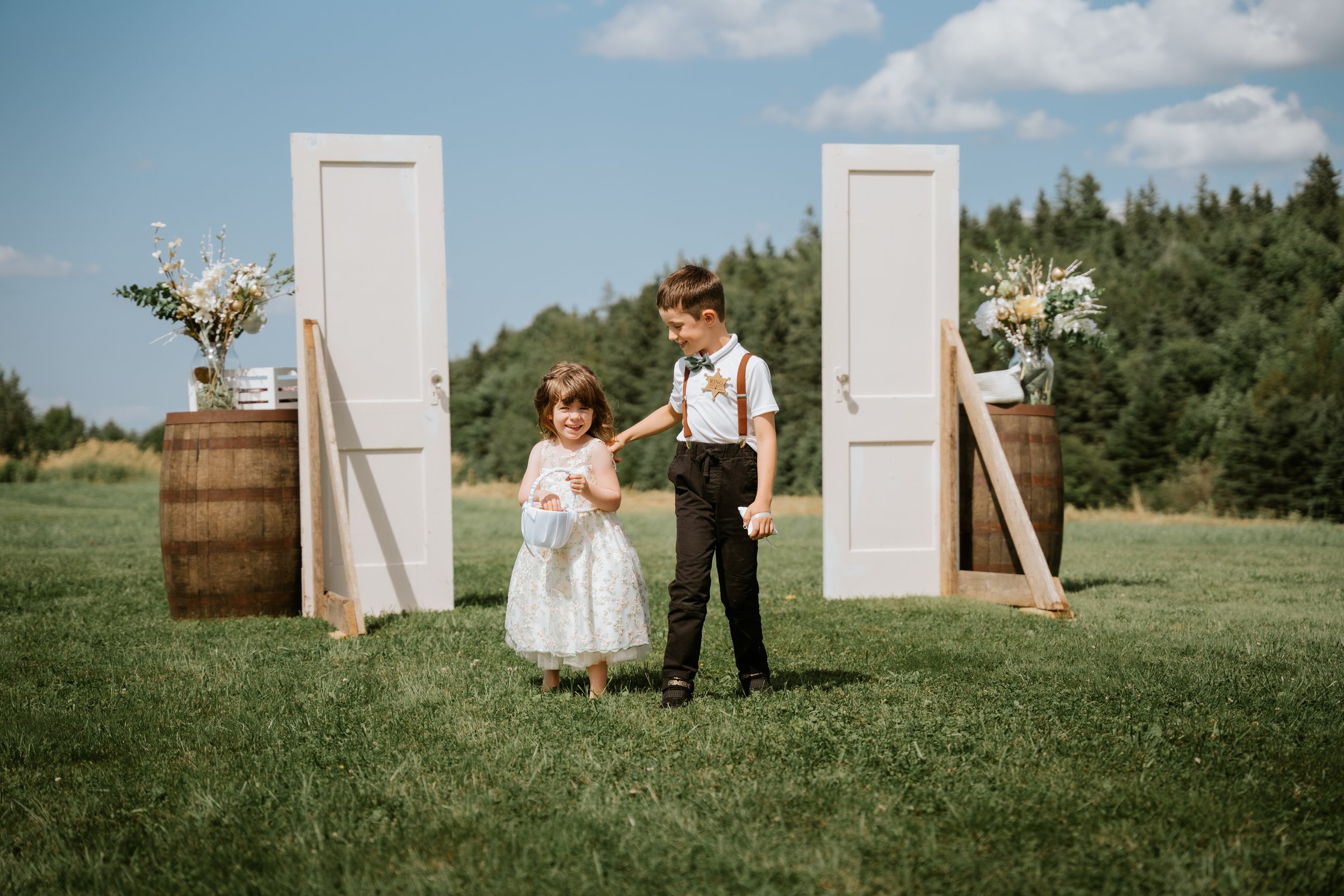 Two children, a girl in a white dress and a boy in black pants and a white shirt with suspenders and a bow tie, walking on a grassy field during a wedding ceremony. They are smiling and holding small baskets. Behind them are two white doors, with flo