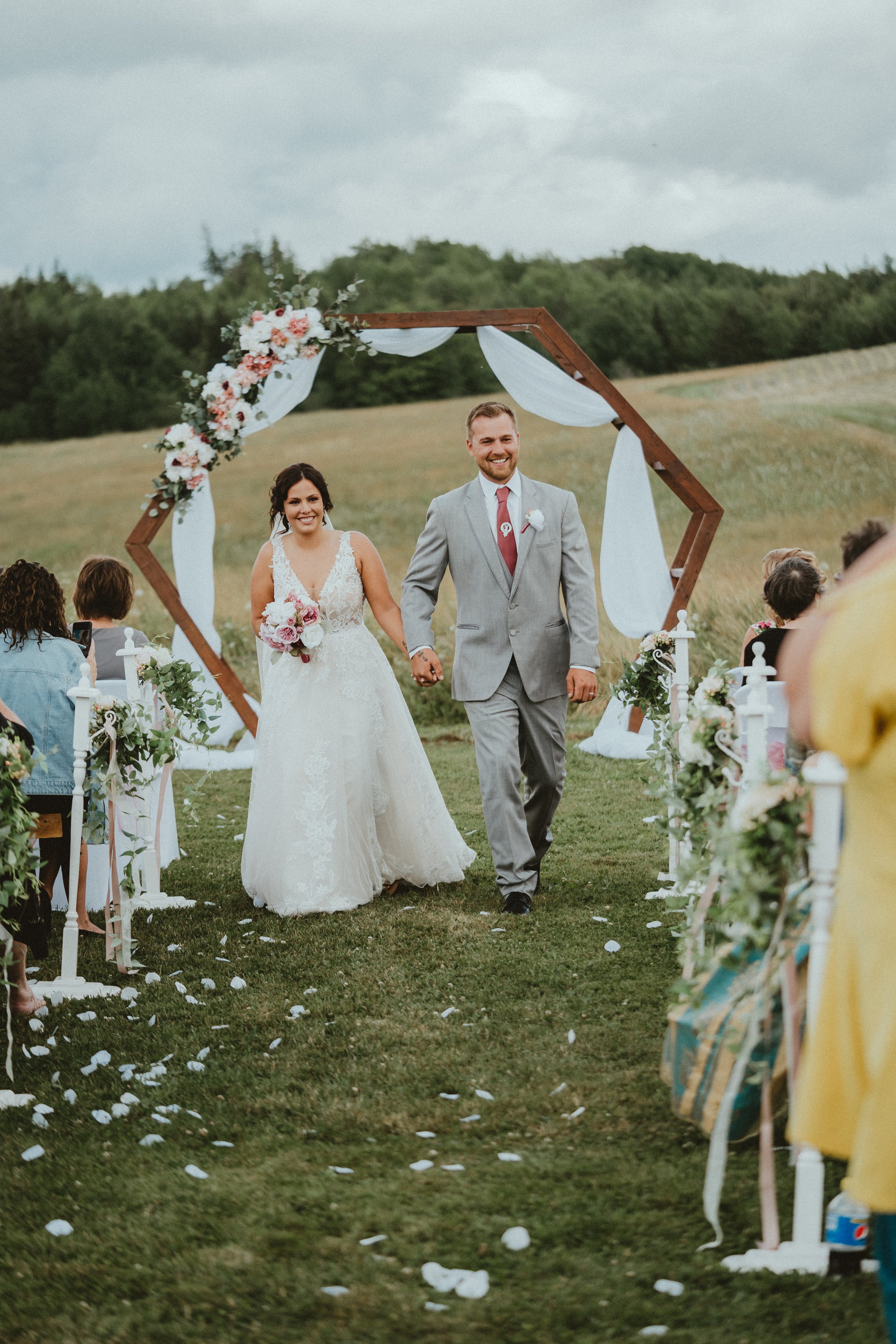 A newlywed couple walks down the aisle outdoors. The bride wears a white lace wedding gown and holds a bouquet, and the groom wears a gray suit with a red tie. They hold hands and smile as they walk under a wooden hexagonal wedding arch decorated wit